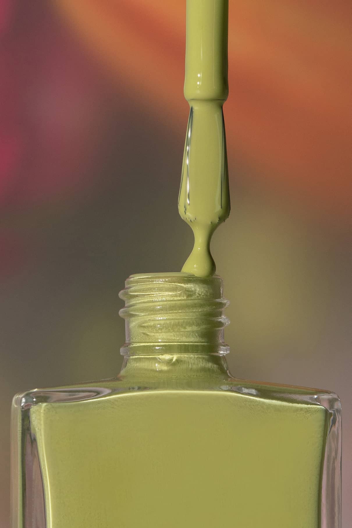 Ein Pinsel trägt den Nagellack gitti no. 196 Nagellack, ein harmonisches Grün, aus einer quadratischen Glasflasche vor einem unscharfen mehrfarbigen Hintergrund.