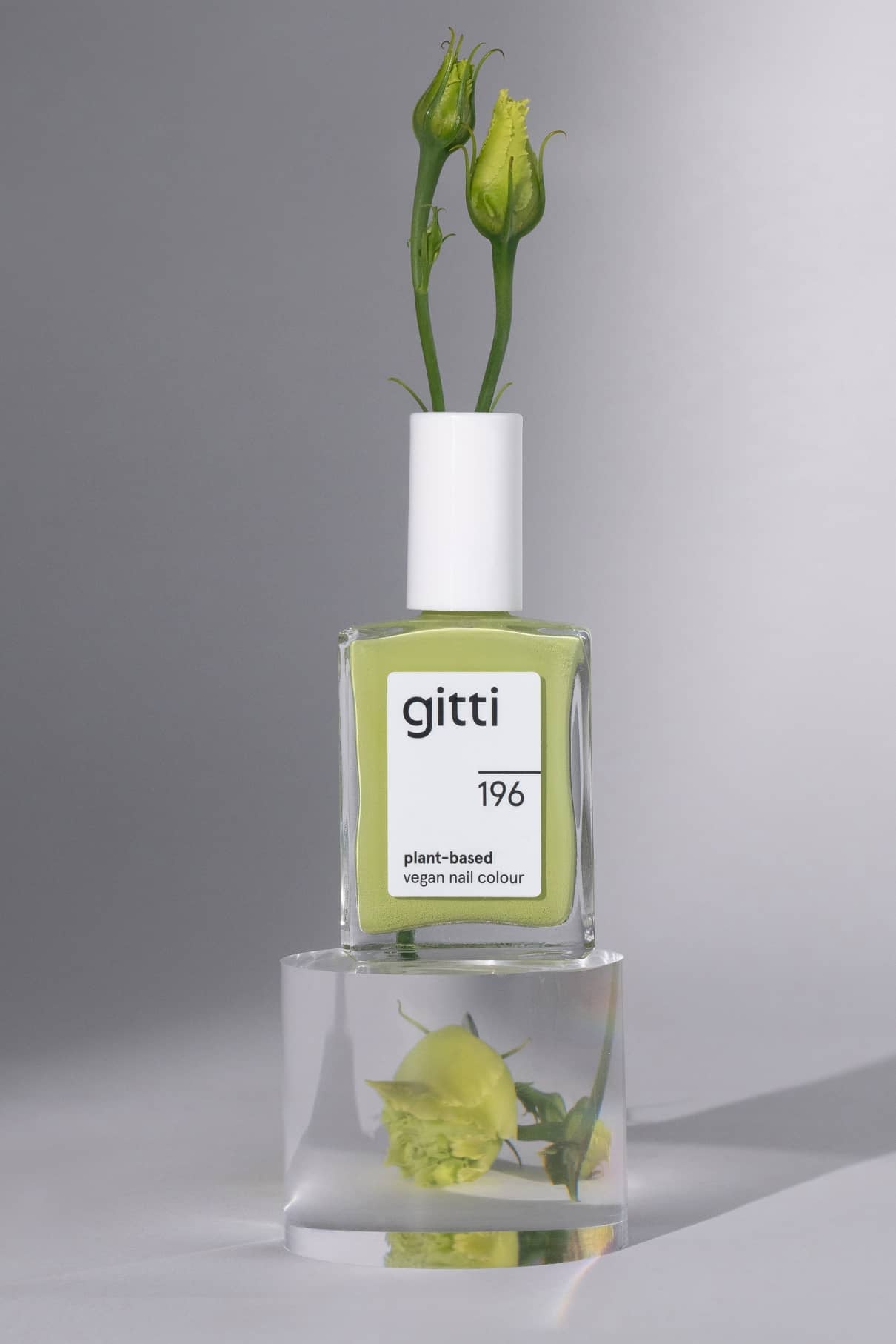 Eine Flasche gitti no. 196 pflanzliche vegane Nagelfarbe, Farbton harmonisches Grün, mit grünen Blütenknospen in der Kappe sitzt auf einem durchsichtigen Block mit einer Blume im Inneren.