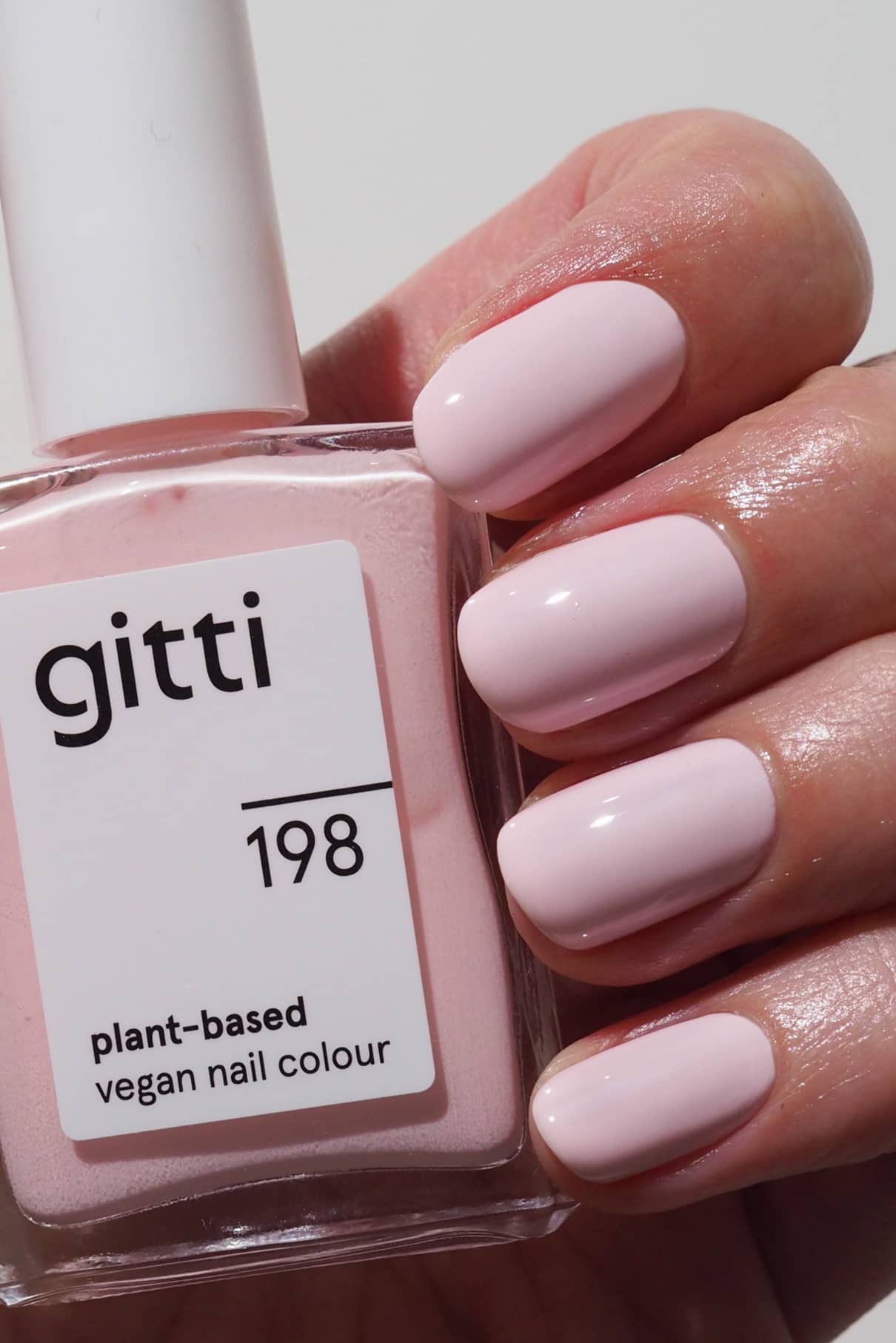 Eine Hand mit kurzen, sauber manikürten und in kühles Rosa lackierten Nägeln hält eine Flasche gitti no. 198 veganen Nagellack auf Pflanzenbasis.