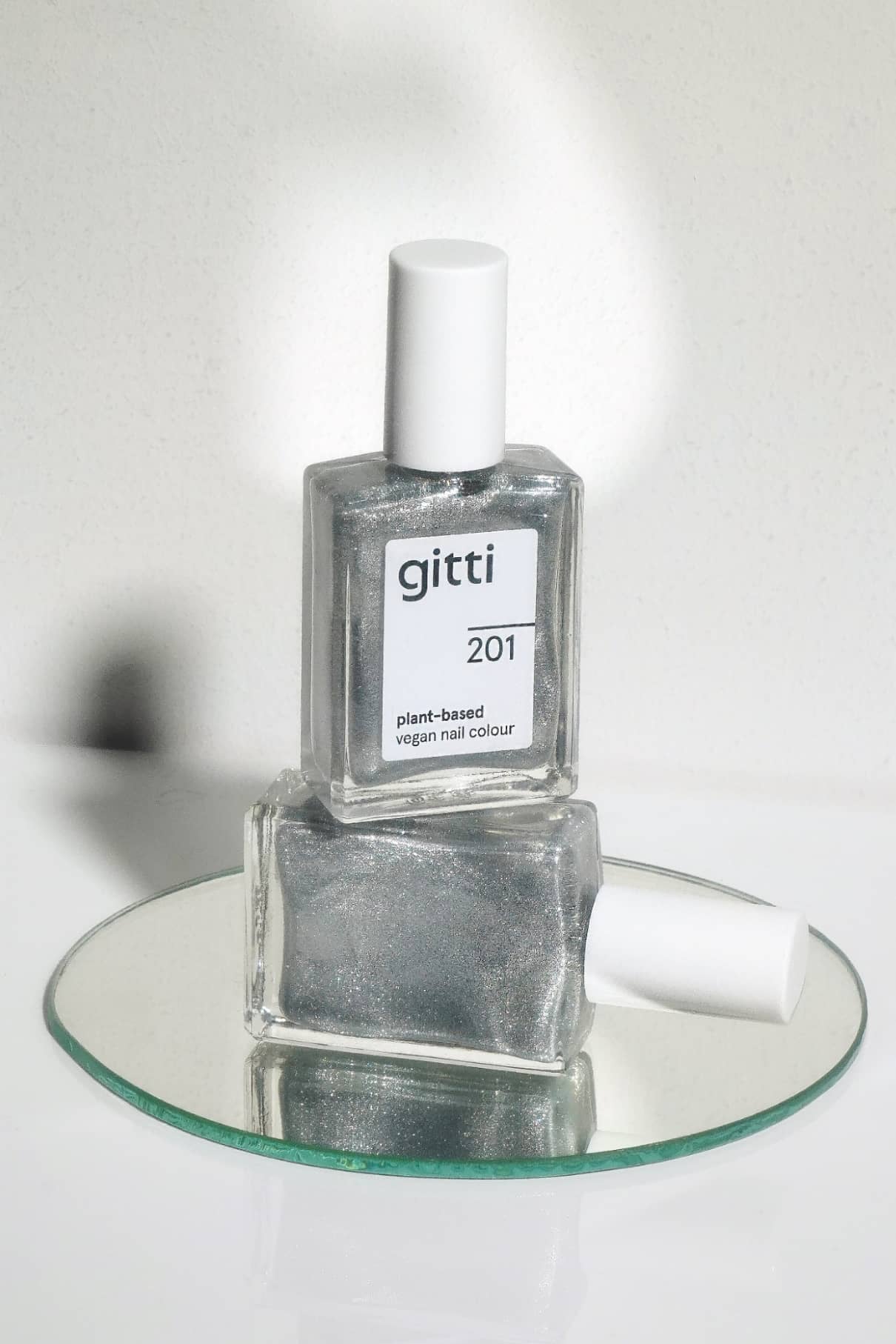 Zwei Flaschen gitti no. 201 pflanzliche vegane Nagelfarbe in kühlem Silber, eine stehend und eine liegend, ausgestellt auf einem runden Spiegel.