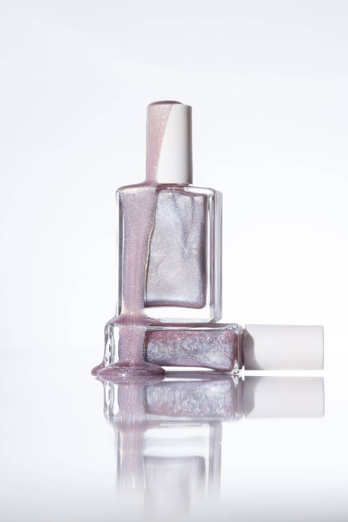 Zwei Flaschen gitti no. 202, ein schimmernder Nagellack in zartem Lavendel: Eine steht, eine liegt, während die mehrfarbige Flüssigkeit von der oberen auf die untere sowie auf die glänzende Oberfläche fließt.