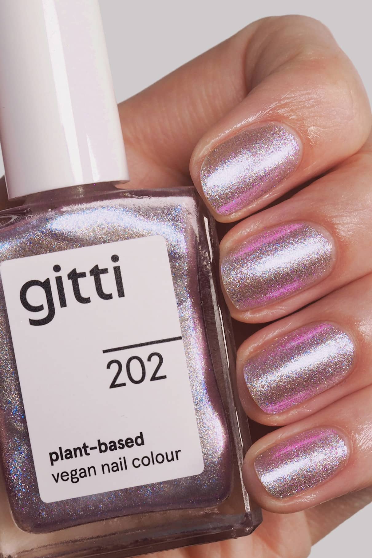 Eine Hand mit kurzen, gepflegten Nägeln, die in einem schimmernden, mehrfarbigen Rosa-Lavendelton lackiert sind, hält eine Flasche mit der Aufschrift "gitti no. 202 Vegane Nagelfarbe auf Pflanzenbasis".
