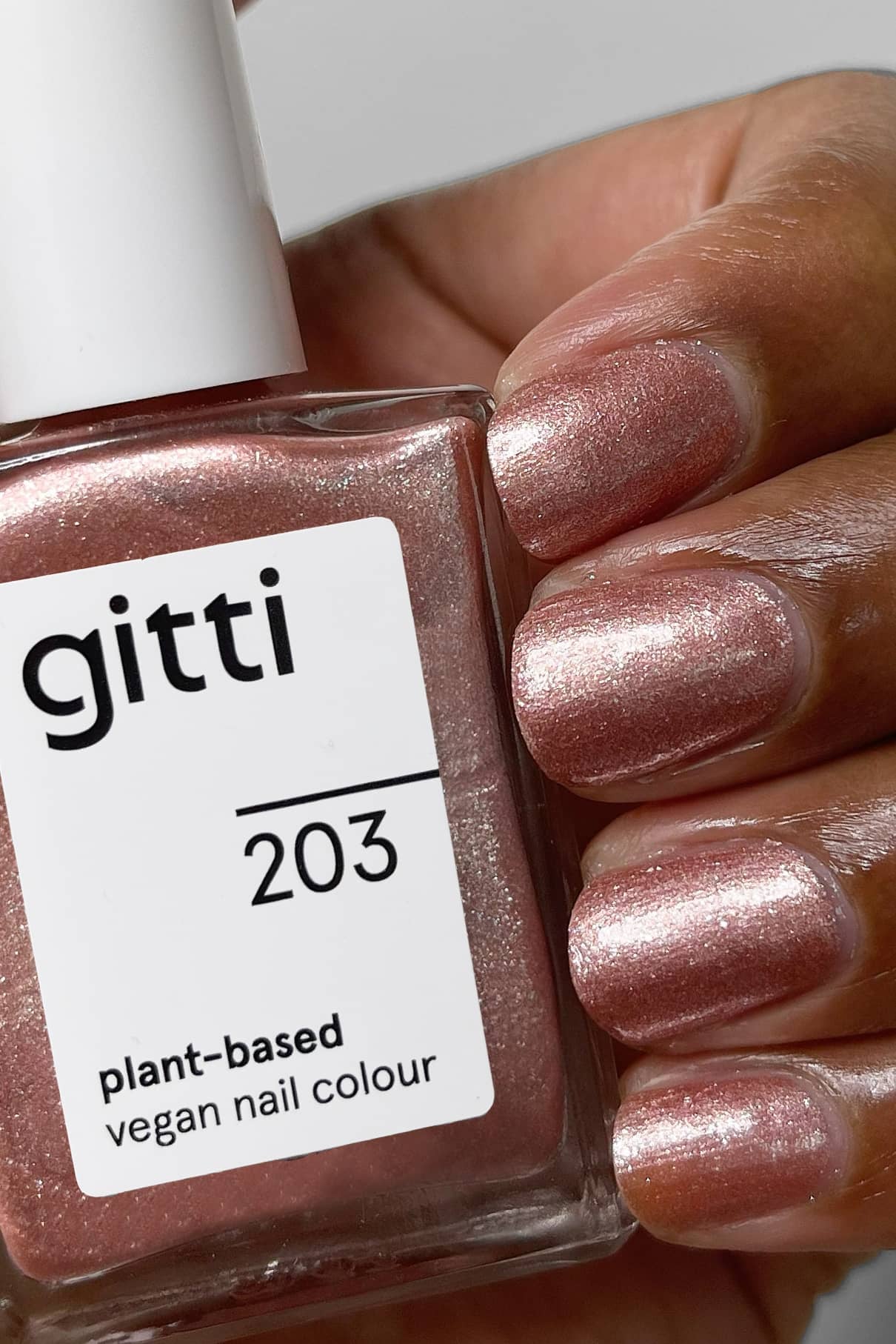Nahaufnahme einer Hand mit schimmerndem rosa, kupferfarbenem Nagellack, die eine passende Flasche mit der Aufschrift gitti no. 203, pflanzliche vegane Nagelfarbe.