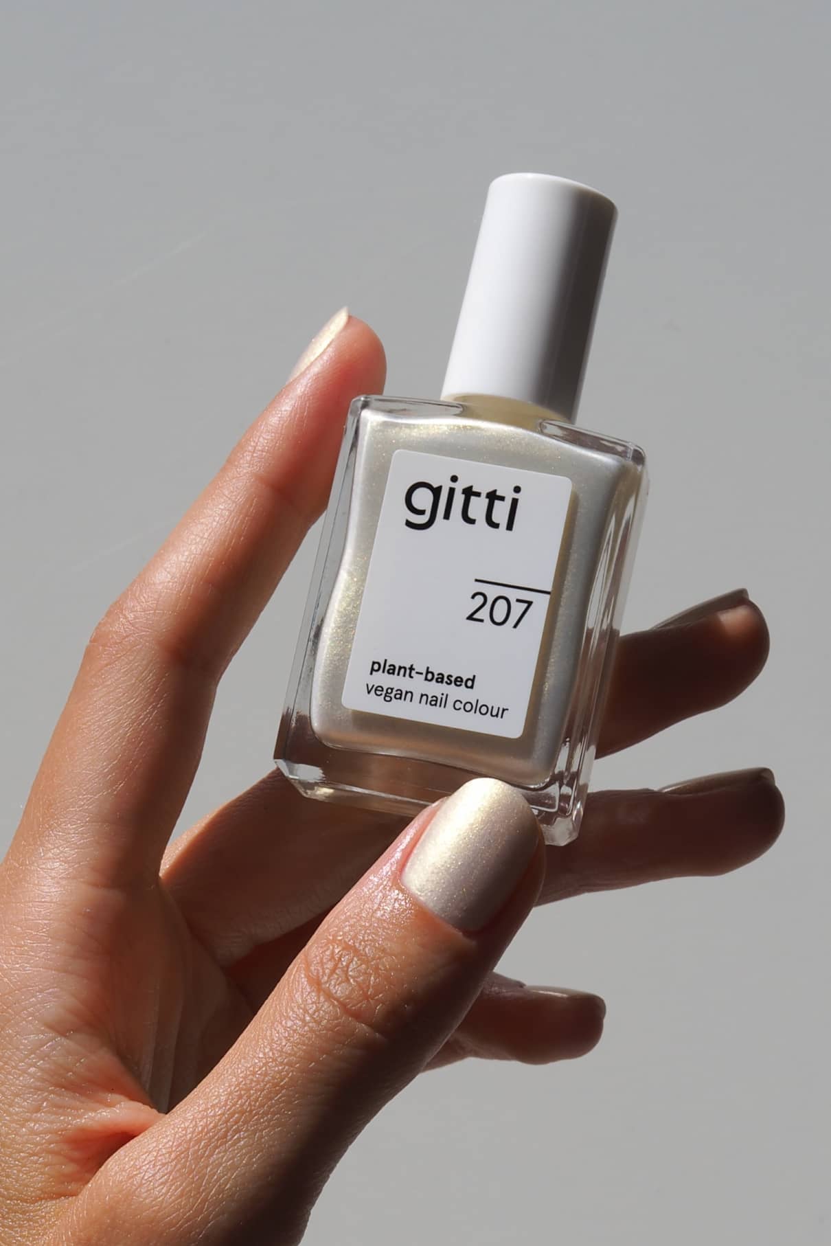 Eine Hand, die eine Flasche gitti no. 207 pflanzliche, vegane Nagelfarbe. Die Nägel sind im gleichen weißen Farbton lackiert, der einen irisierenden Schimmer aufweist.