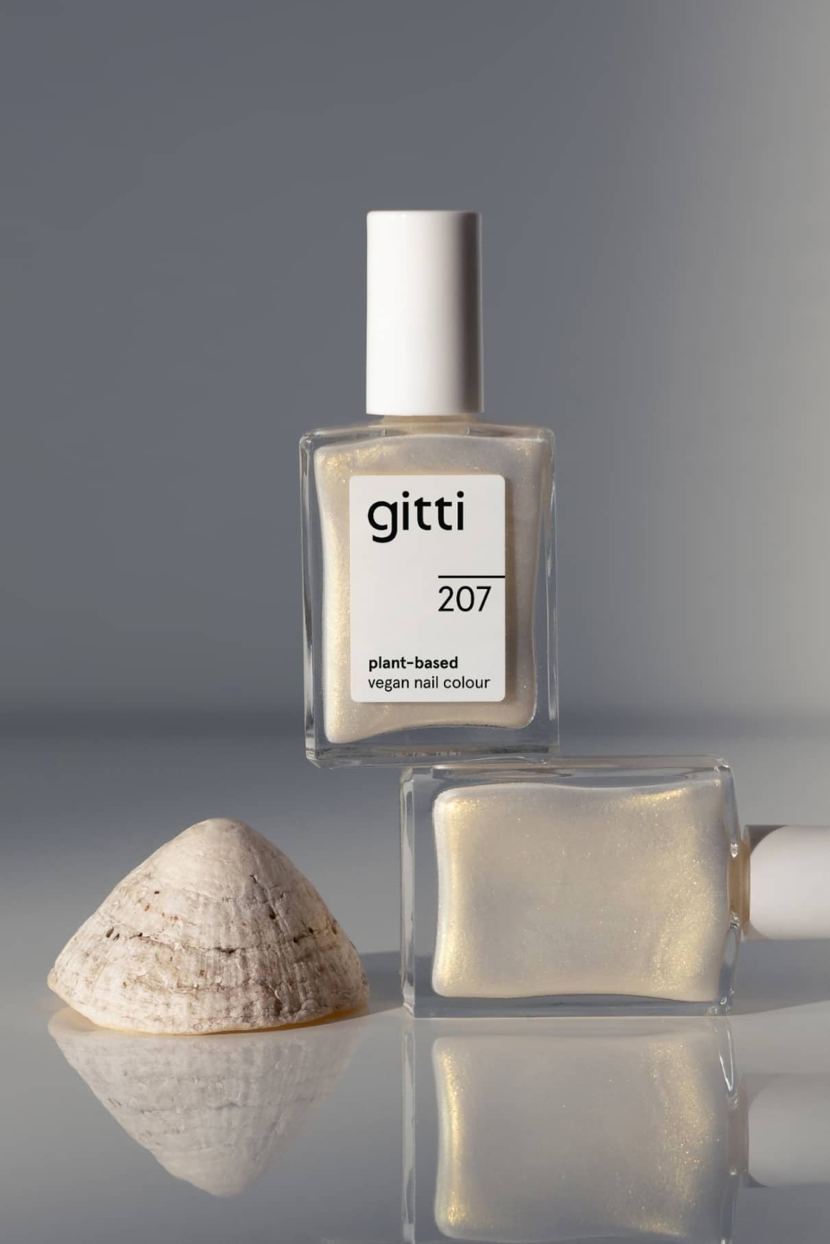 Zwei Flaschen gitti no. 207 pflanzliche vegane Nagelfarbe in einem zarten Weißton mit irisierendem Schimmer stehen neben einer Muschel auf einer spiegelnden Oberfläche.