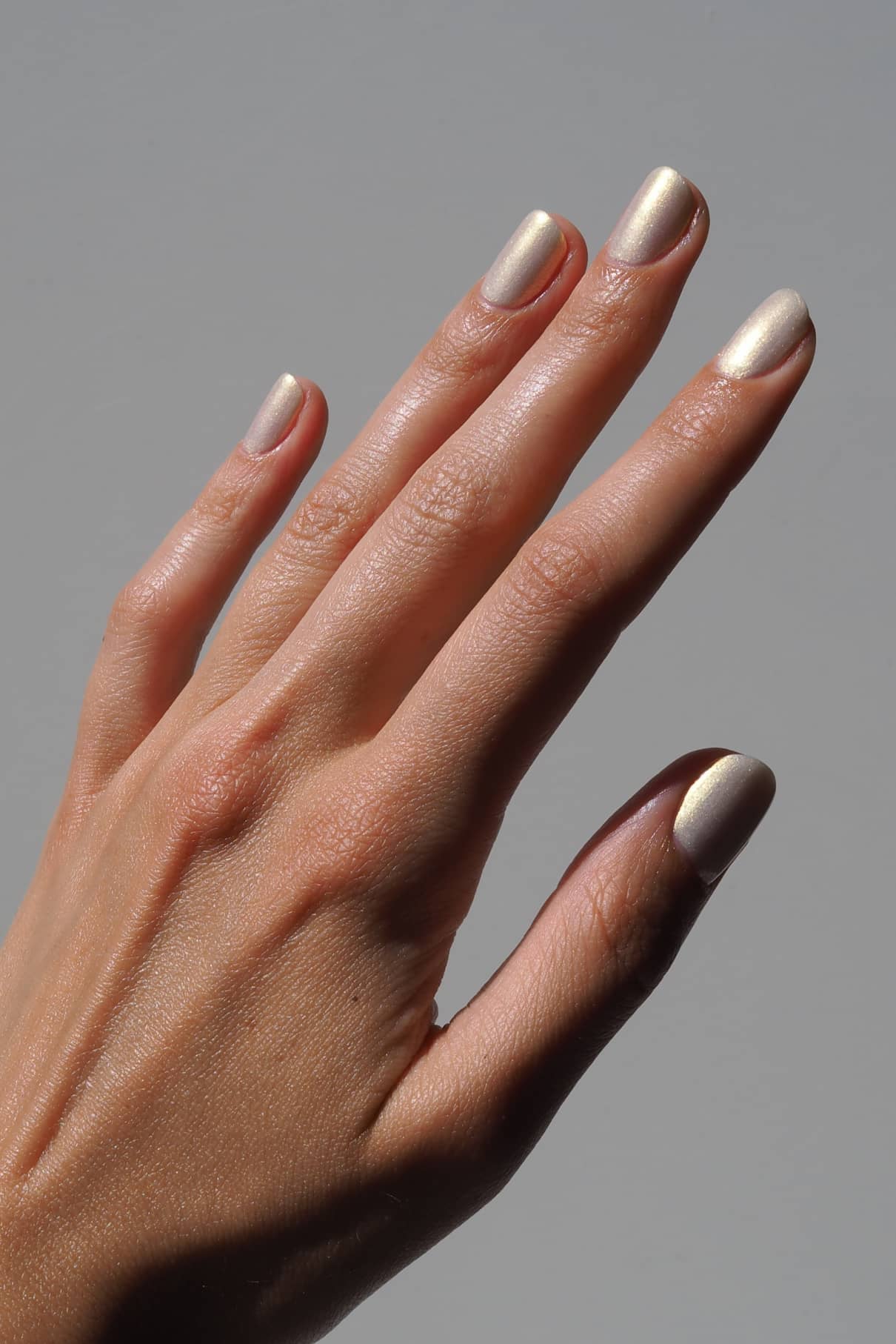 Eine Hand mit sauber manikürten Nägeln, lackiert mit gitti no. 207, einem metallischen gold-bläulichen Lack mit irisierendem Schimmer, vor einem schlichten grauen Hintergrund.