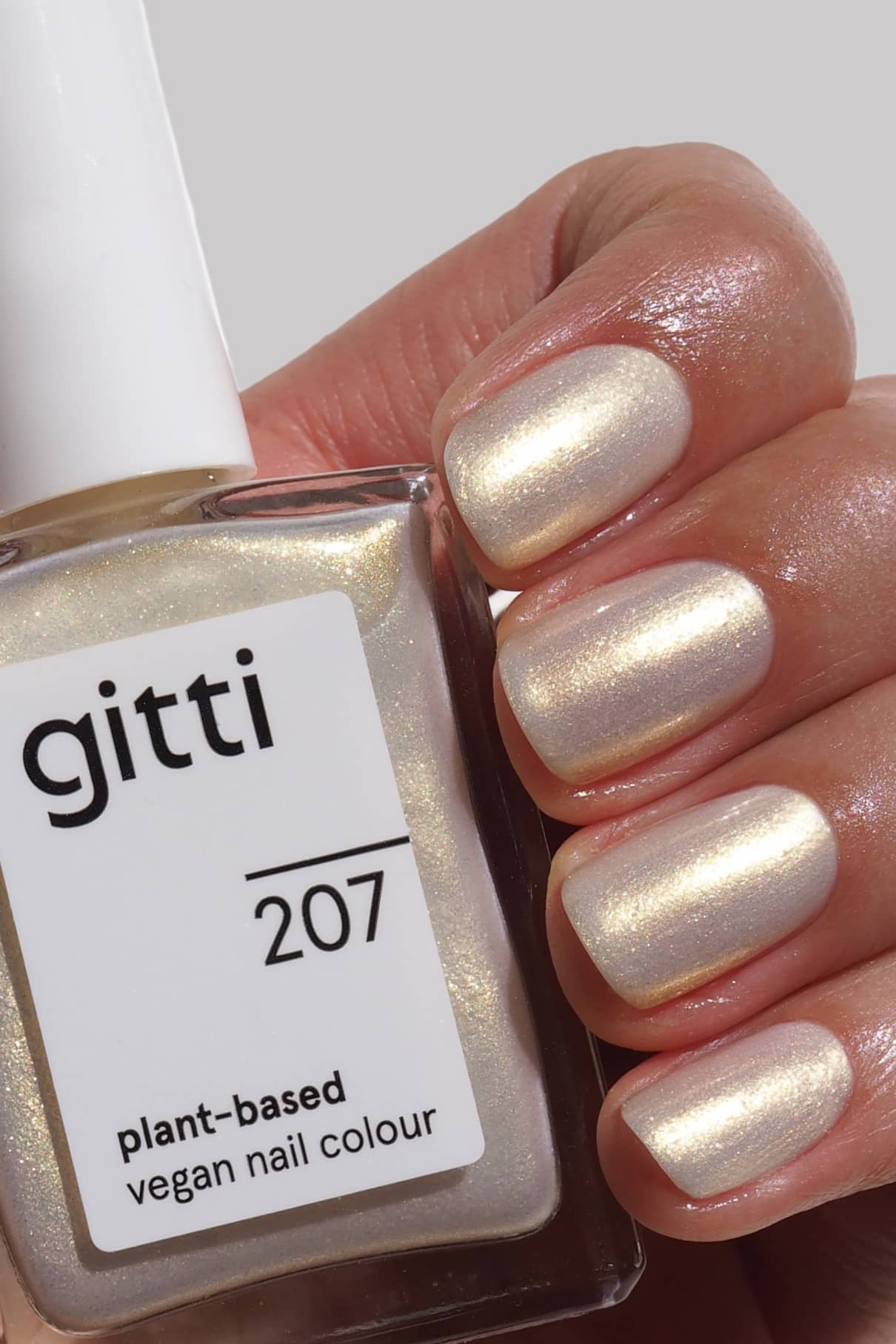 Eine Hand mit kurzen, sauber manikürten Nägeln in weißem, gold-blau irisierendem Schimmer hält eine Flasche mit gitti no. 207 pflanzlicher veganer Nagelfarbe.