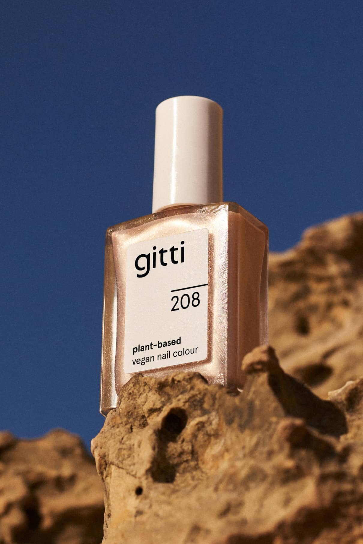 Ein Fläschchen mit gitti no. 208 pflanzliche, vegane Nagelfarbe in glitzerndem Gold sitzt auf einem rauen Felsen vor einem klaren blauen Himmel.