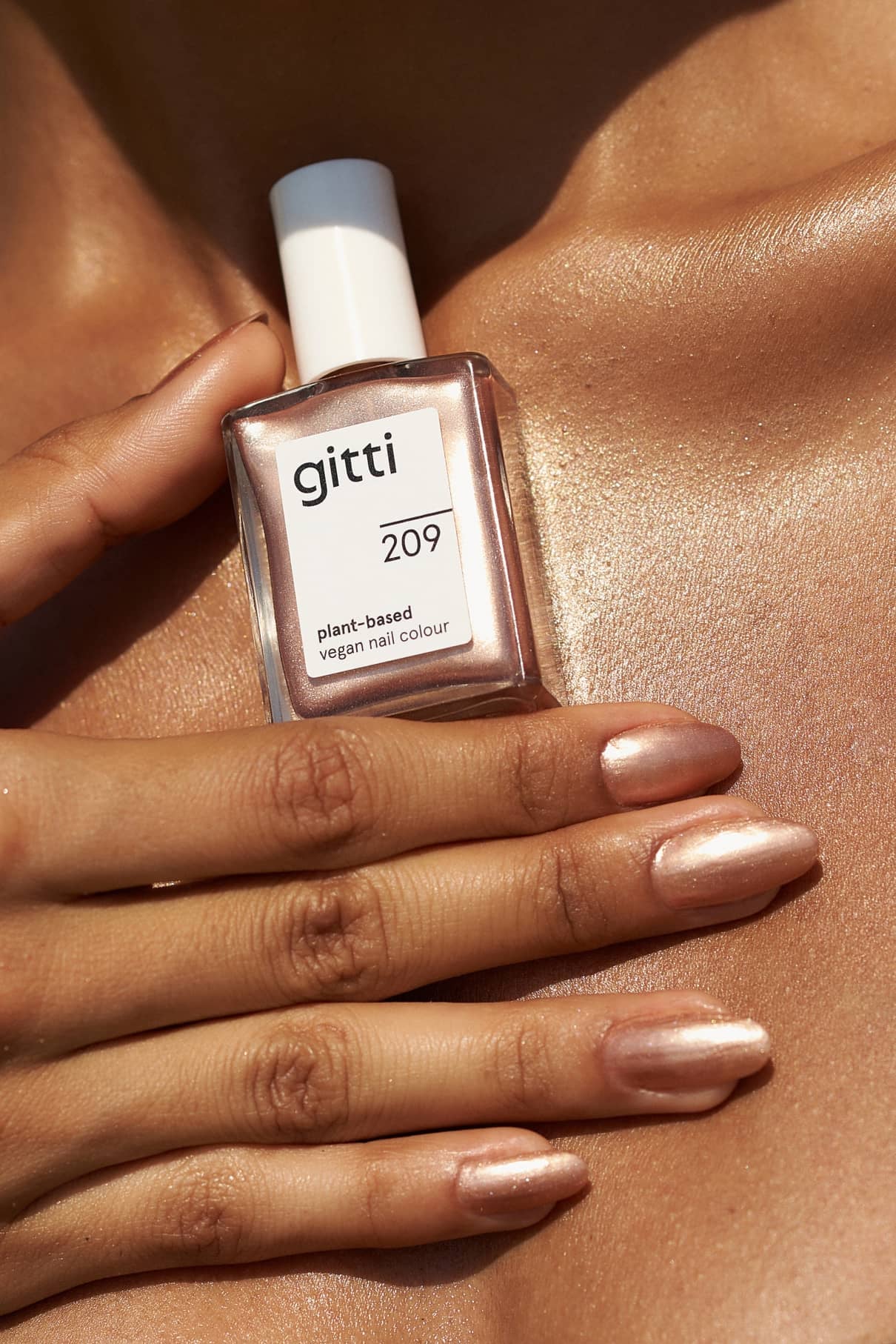 Eine Hand mit metallischem Nude-Nagellack in Bronzeton hält eine Flasche gitti no. 209 pflanzlicher, veganer Nagellack auf gebräunter, leuchtender Haut.