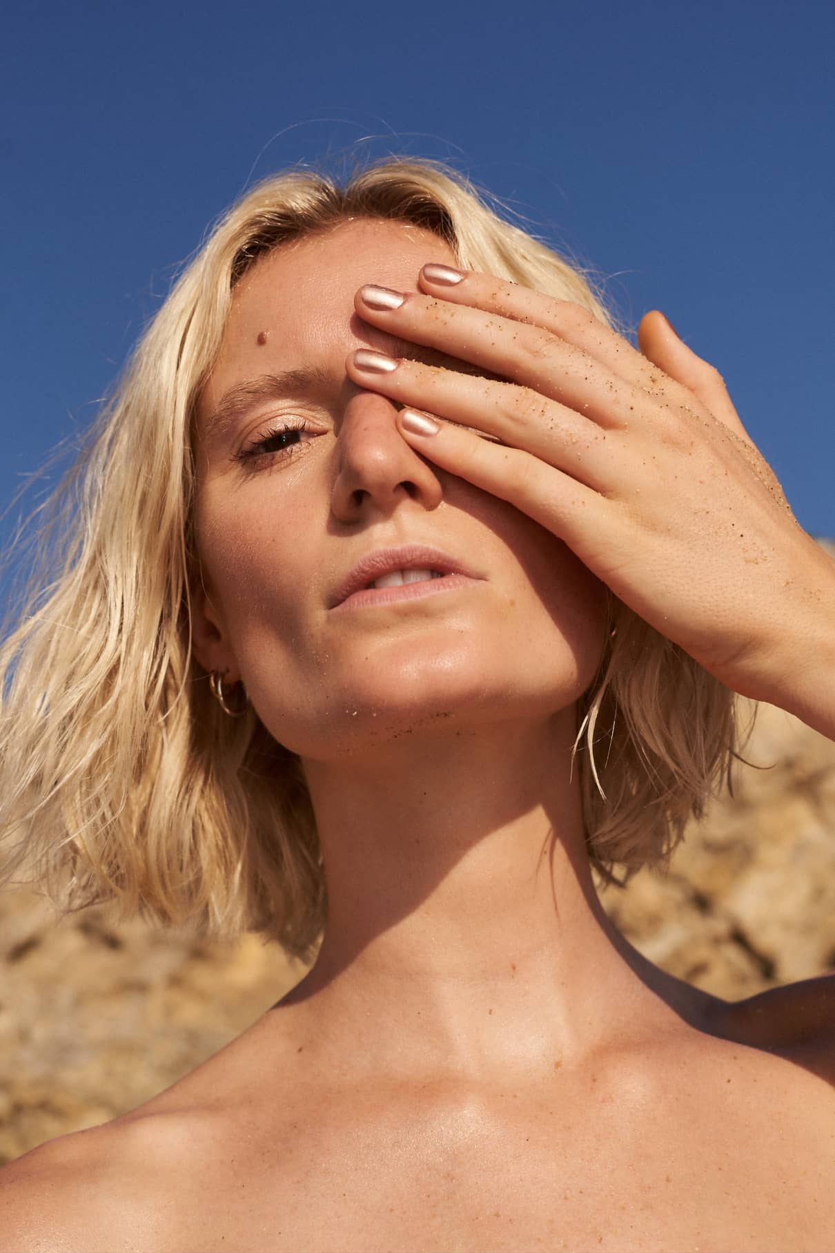 Eine Person mit kurzen blonden Haaren und bronzetonfarbener Haut bedeckt ein Auge mit einer sandfarbenen Hand und zeigt die Gitti Nr. 209, während sie im Freien unter einem klaren blauen Himmel steht.