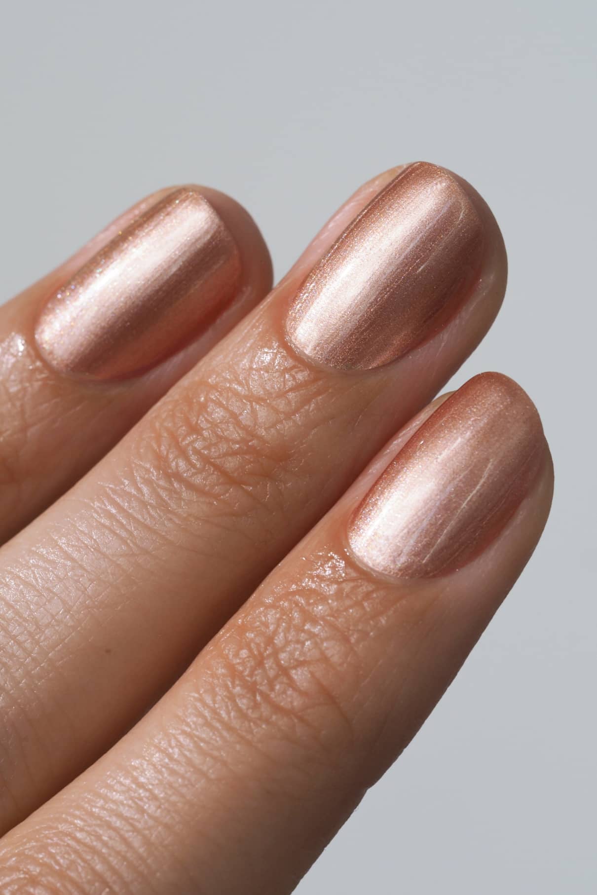 Nahaufnahme von drei Fingern mit kurzen Nägeln, lackiert mit metallisch glänzendem, roségoldenem gitti no. 209 Nagellack mit einem Hauch von Bronzeton vor einem schlichten Hintergrund.