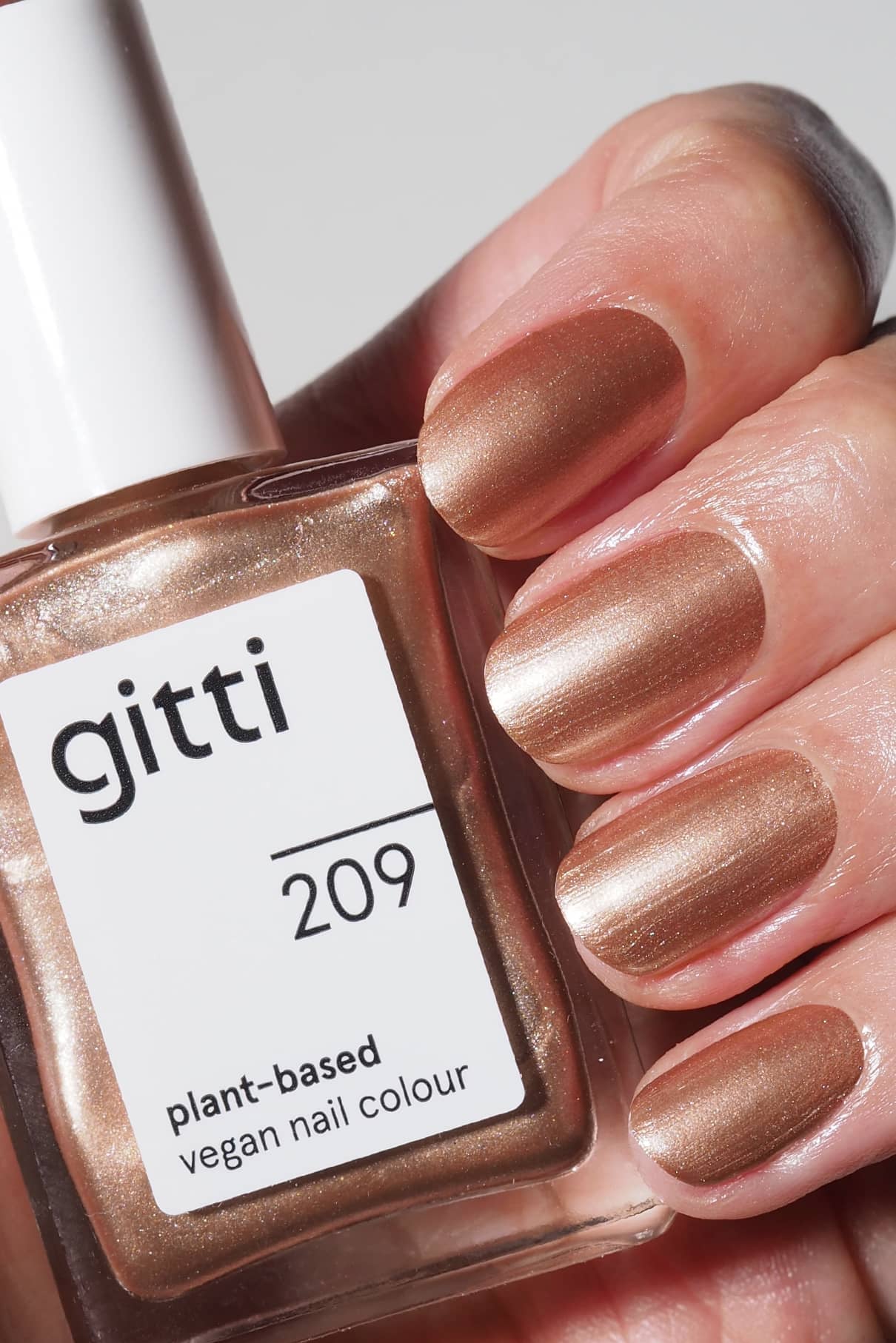 Eine Hand mit kurzen Nägeln, die in einem satten Bronzeton-Metallic-Ton lackiert sind, hält ein Fläschchen mit gitti no. 209 pflanzlicher, veganer Nagellack.