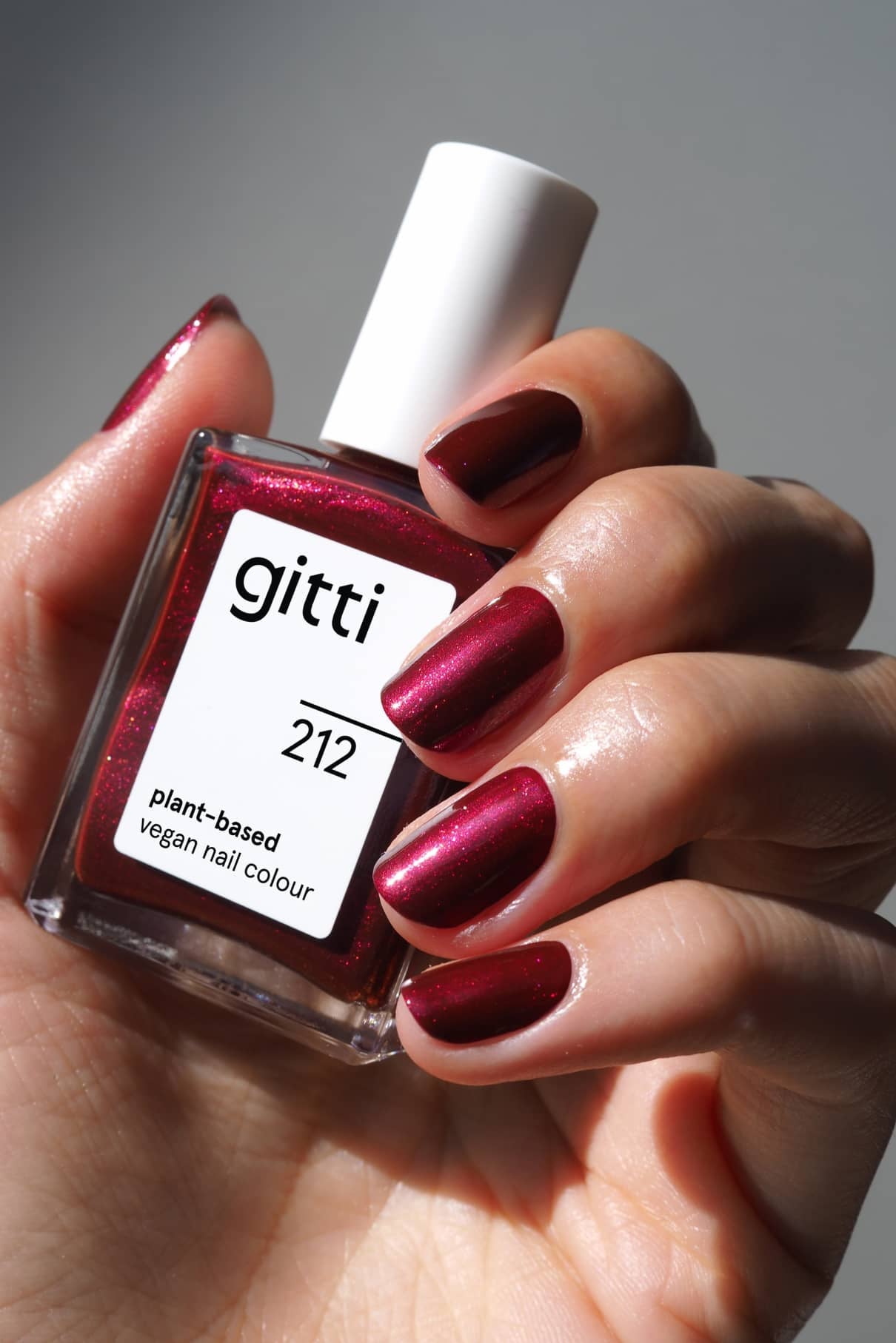 Eine Hand hält eine Flasche mit gitti no. 212, einem pflanzlichen, veganen Nagellack; die Nägel zeigen einen glänzenden, beerenroten Farbton mit einem subtilen metallischen Schimmer.
