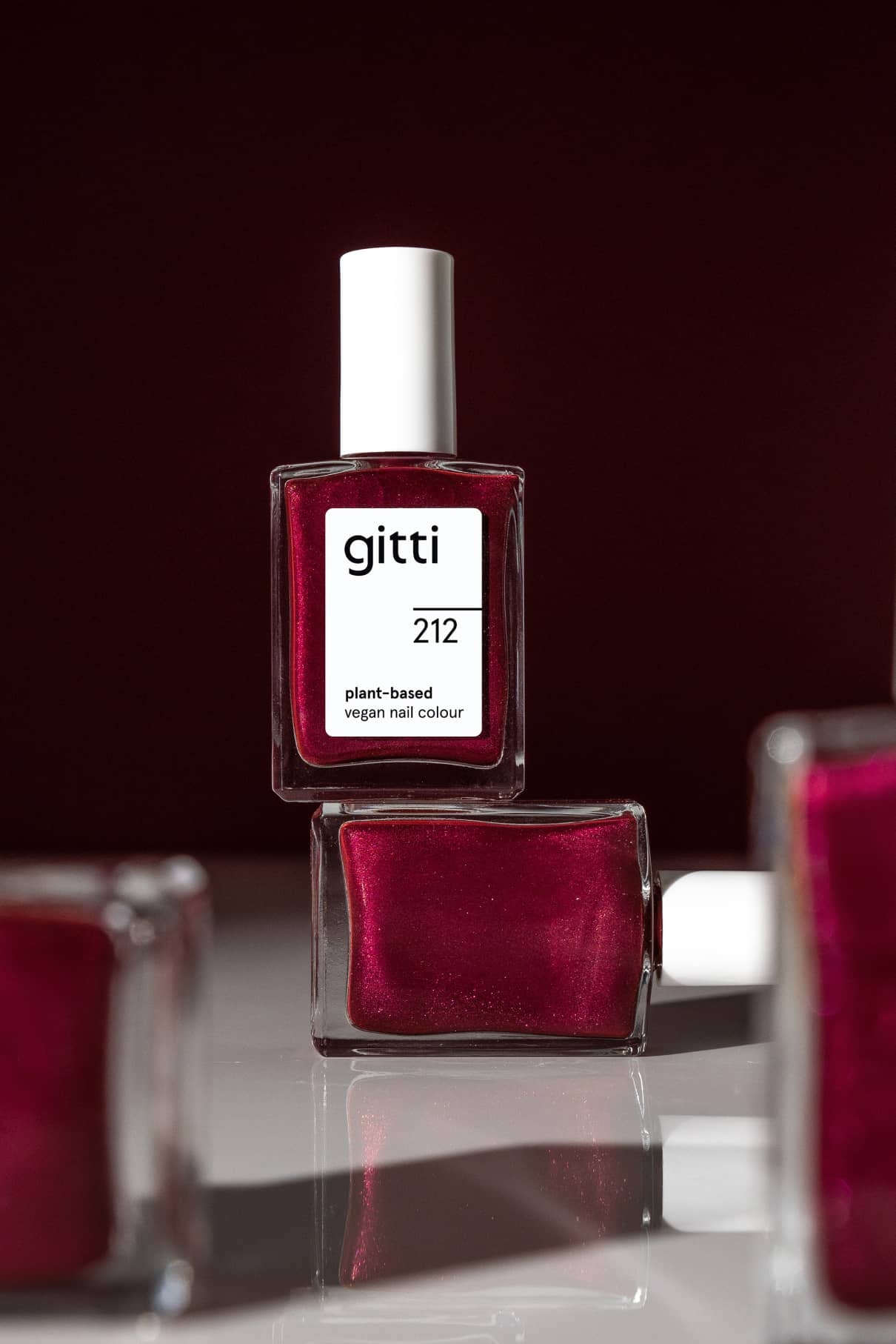 Drei Flaschen von gitti no. 212, ein veganer Nagellack auf pflanzlicher Basis in bierigem Rot mit metallischem Schimmer, sind ausgestellt - eine aufrecht über der anderen und eine teilweise sichtbar im Vordergrund.
