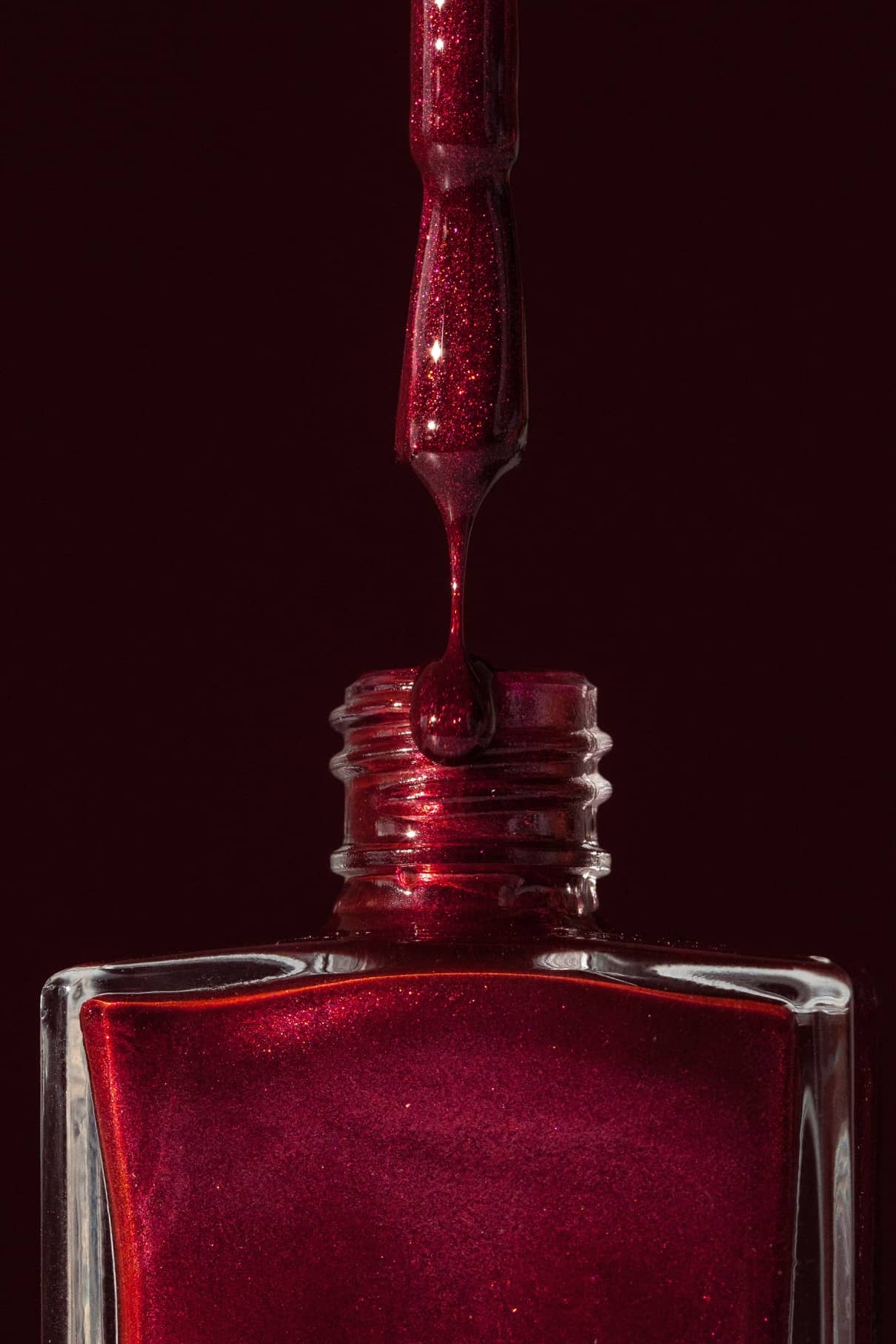 Eine Nahaufnahme eines Nagellackpinsels mit gitti no. 212 Metallisch schimmernder roter Lack, der in eine passende beerenrote Flasche vor einem dunkelroten Hintergrund tropft.