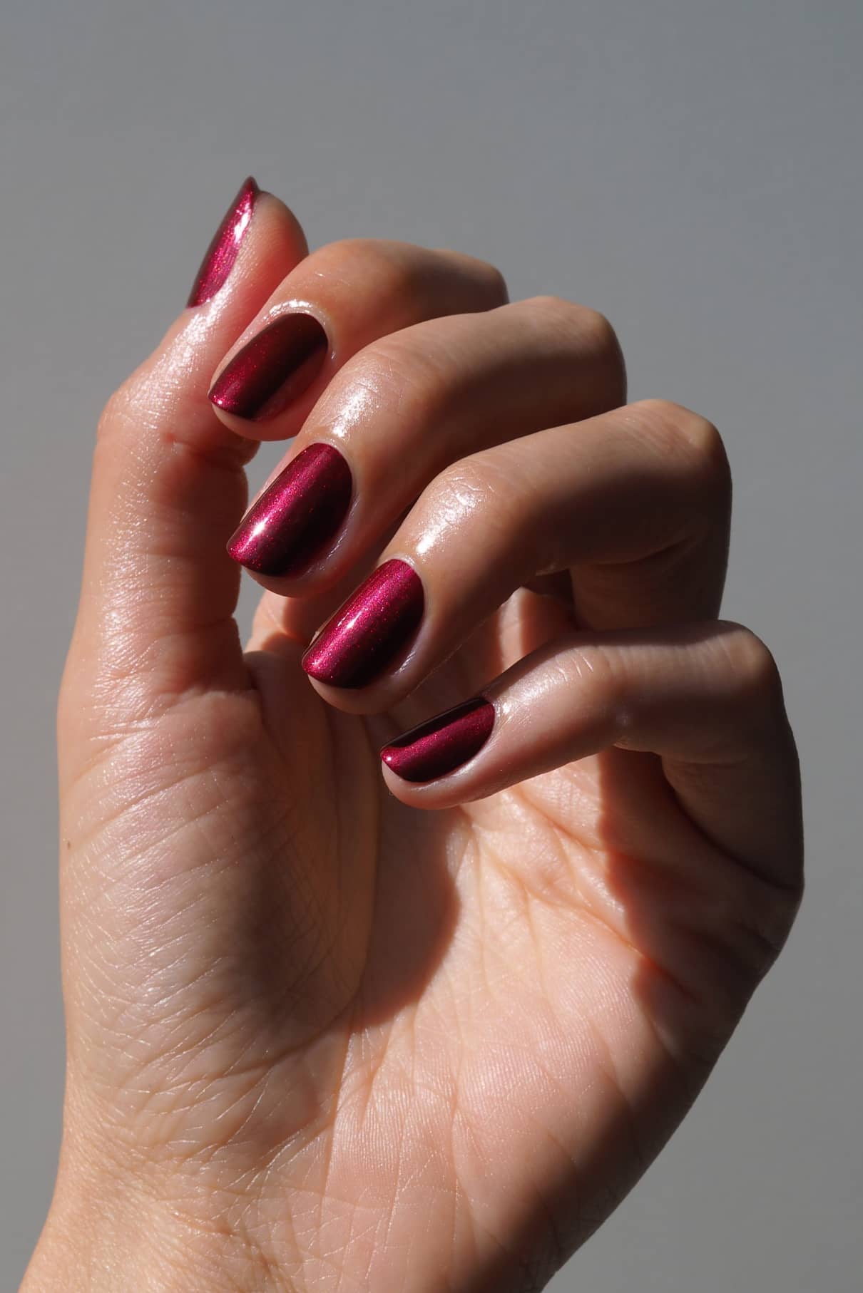Eine Hand mit leicht gekrümmten Fingern zeigt Nägel, die in gitti no. 212, einem beerigen Rot mit Metallic-Schimmer, vor einem neutralen Hintergrund.
