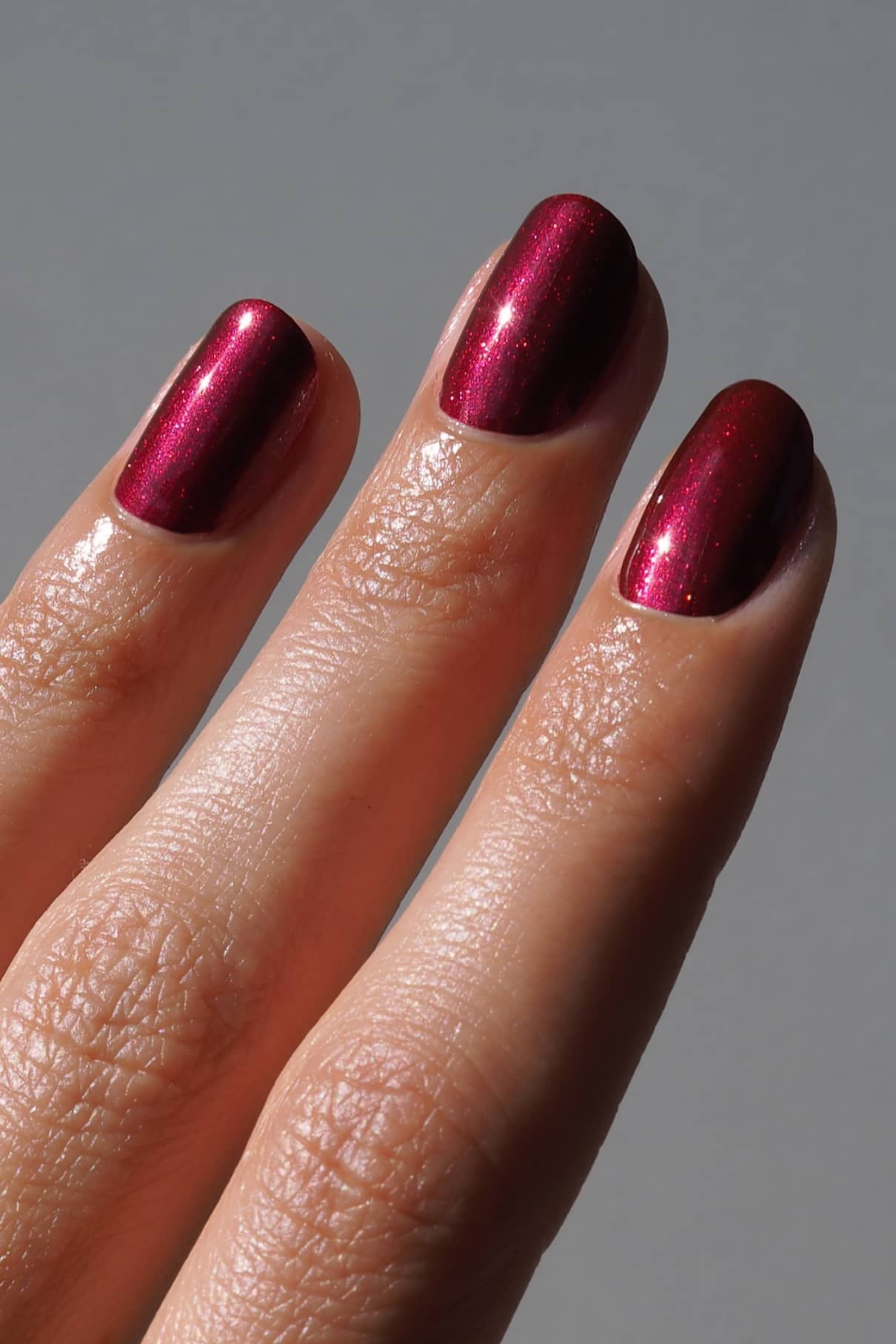 Eine Nahaufnahme von drei Fingern mit kurzen, glänzenden Nägeln, lackiert in gitti no. 212, einem beerigen Rot-Ton mit Metallic-Schimmer, vor einem schlichten grauen Hintergrund.
