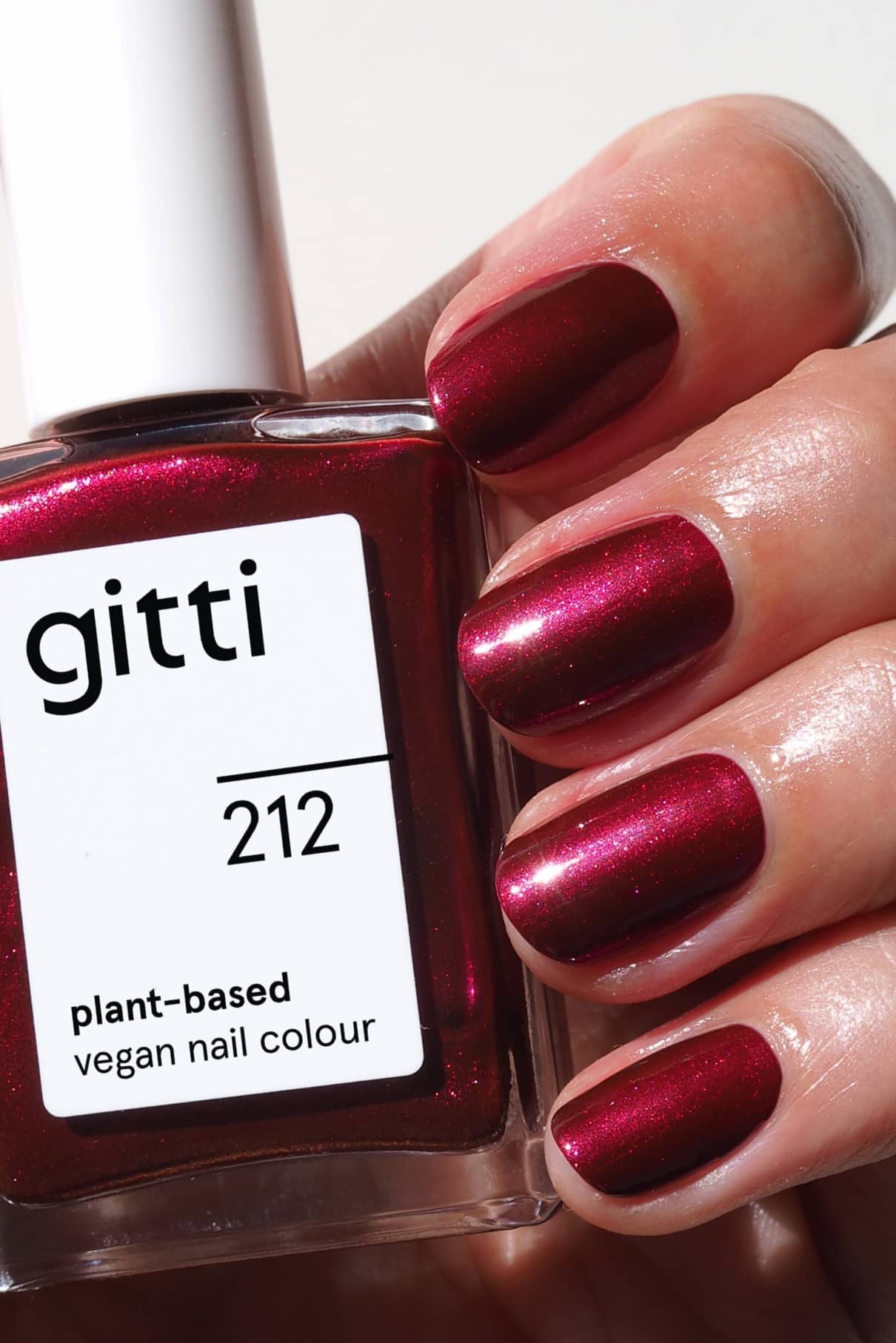 Eine Hand mit kurzen Nägeln, die in einem Beerenrot mit metallischem Schimmer lackiert sind, hält eine Flasche gitti no. 212 vegane Nagelfarbe auf Pflanzenbasis.