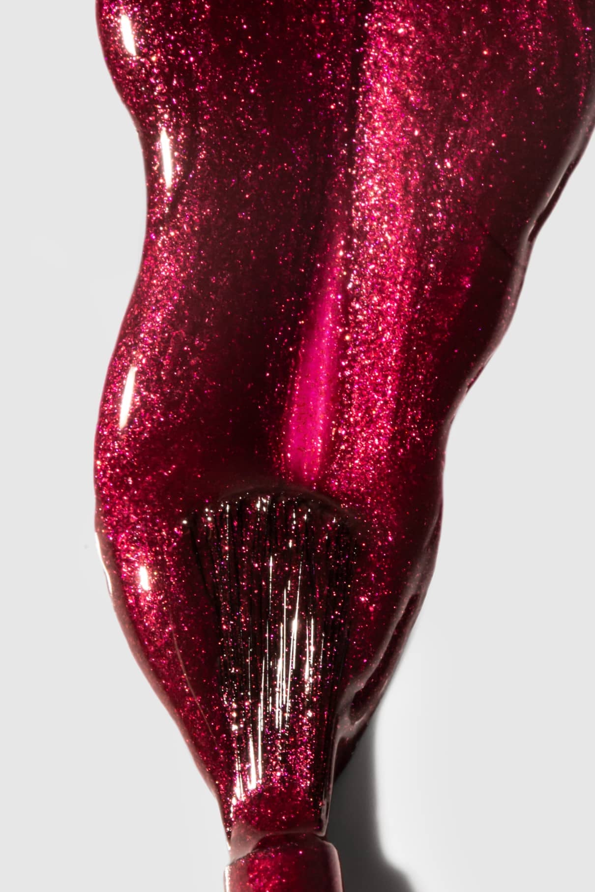 Nahaufnahme eines Pinsels, mit dem gitti no. 212, ein glänzender, metallisch schimmernder, beerenroter Nagellack, auf einer weißen Fläche.