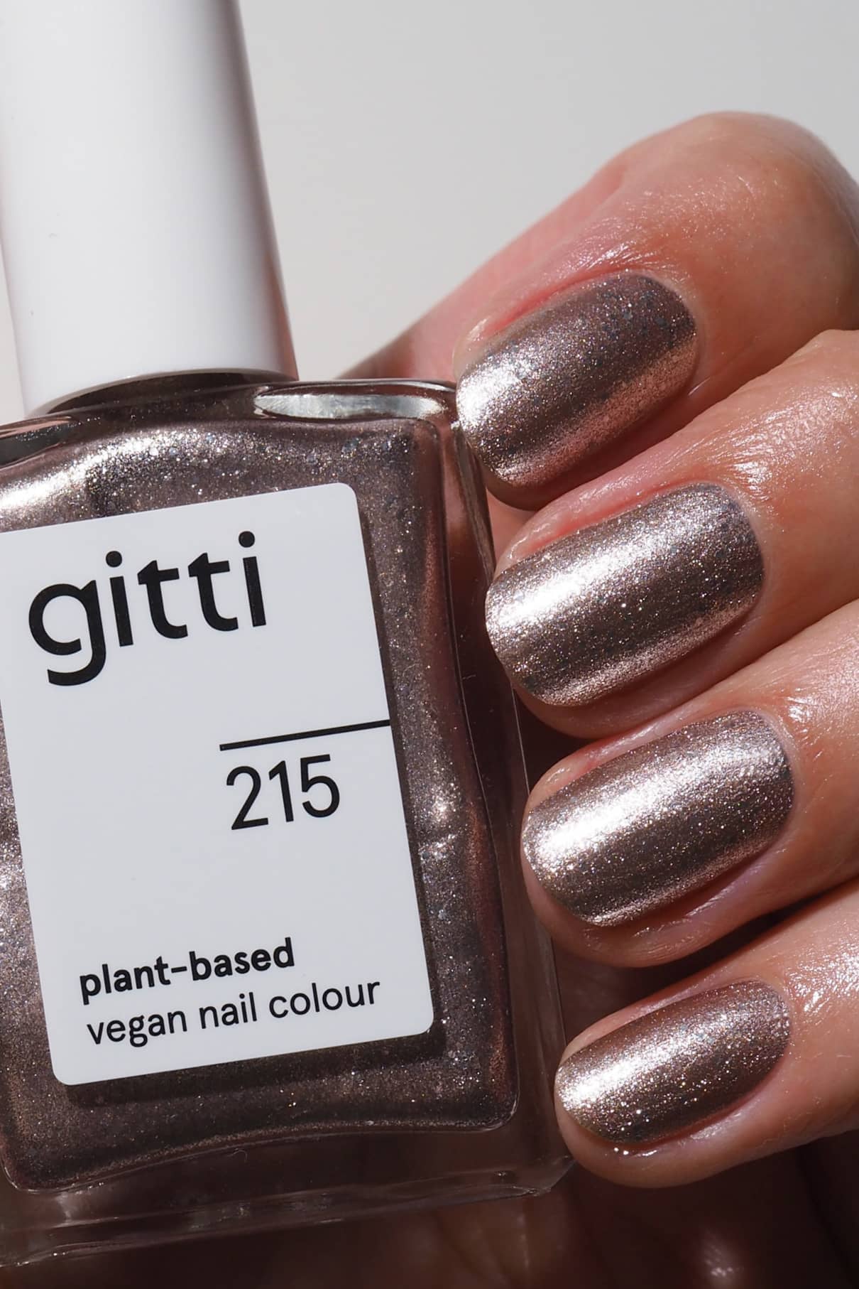 Eine Hand mit metallisch rosa schimmernden Nägeln hält eine Flasche mit der Aufschrift gitti no. 215, eine vegane Nagelfarbe auf Pflanzenbasis.