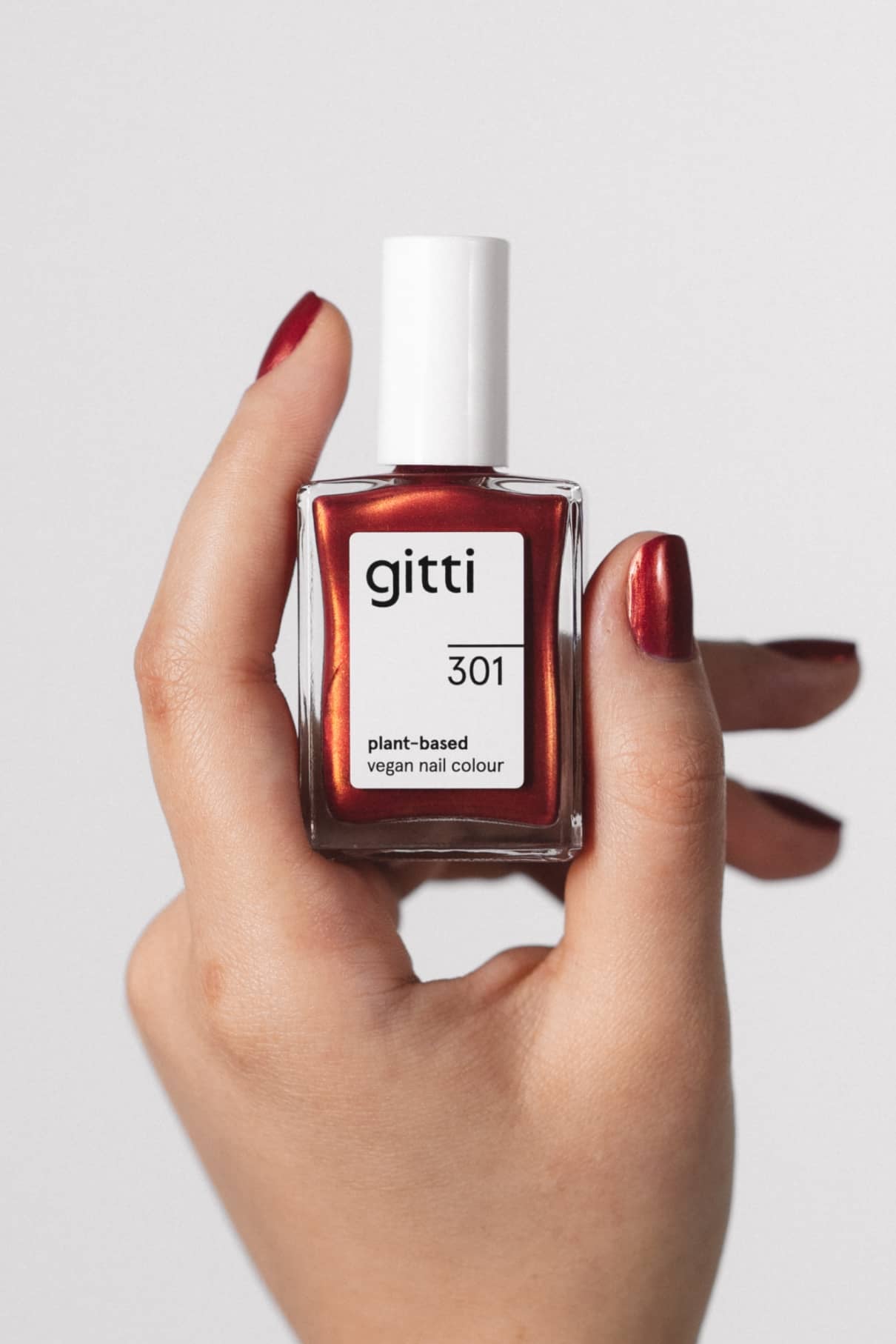 Eine Hand mit lackierten Nägeln hält eine Flasche mit gitti no. 301, einer veganen Nagelfarbe auf Pflanzenbasis in Metallic-Rot, vor einem schlichten Hintergrund.