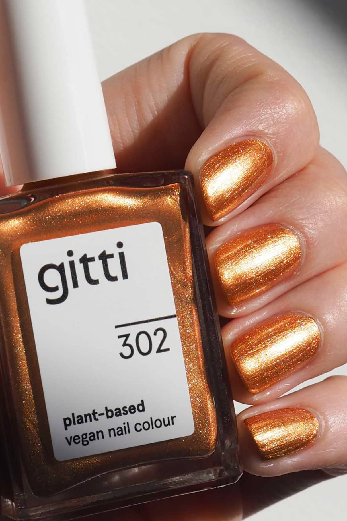 Eine Hand mit metallischem Bronzeton-Nagellack hält eine Flasche mit der Aufschrift gitti no. 302 pflanzlich-vegane Nagelfarbe.