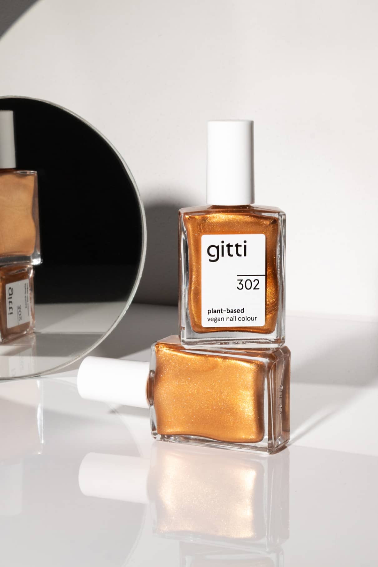 Zwei Flaschen von gitti no. 302, ein goldfarbener veganer Nagellack auf pflanzlicher Basis in Metallic-Bronzeton - eine aufrecht, eine liegend - sind mit einem runden Spiegel ausgestellt, der einen Teil der Szene reflektiert.