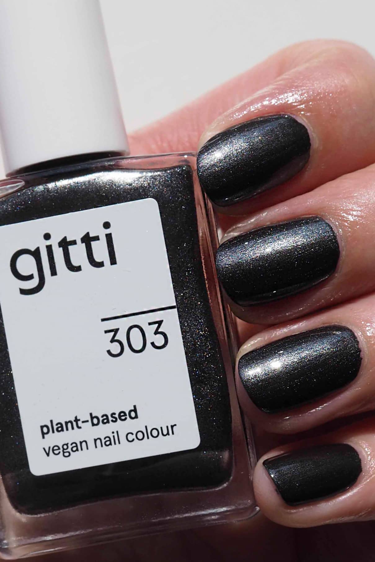 Eine Hand mit vier in Schwarz mit silbrigem Metallic-Finish lackierten Nägeln hält eine Flasche "gitti no. 303, vegane Nagelfarbe auf Pflanzenbasis".