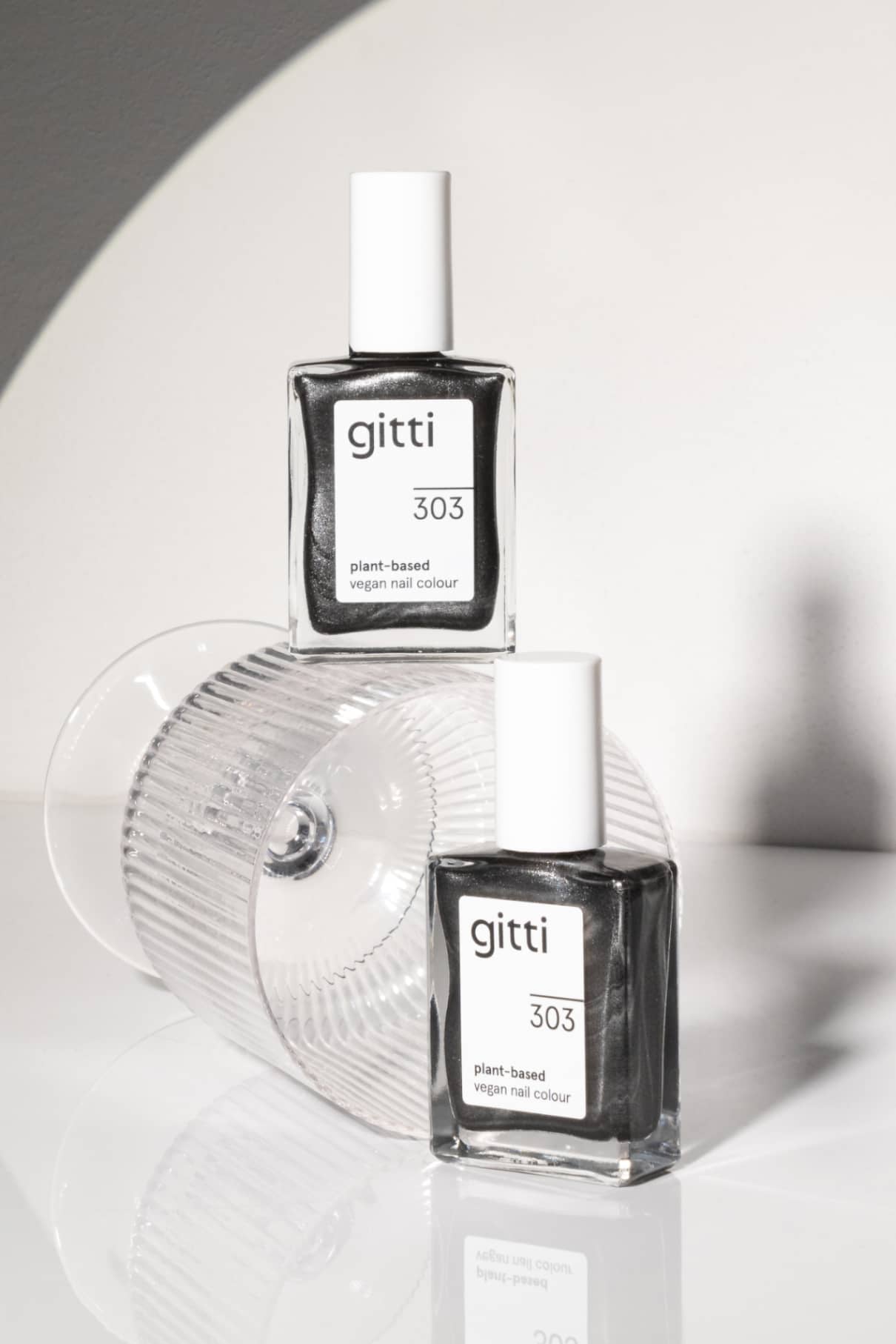 Zwei Flaschen von gitti no. 303 pflanzliche vegane Nagelfarbe mit silbrigem Metallic-Finish sind abgebildet: eine aufrecht auf einem umgestürzten Klarglas, die andere dagegen gelehnt.