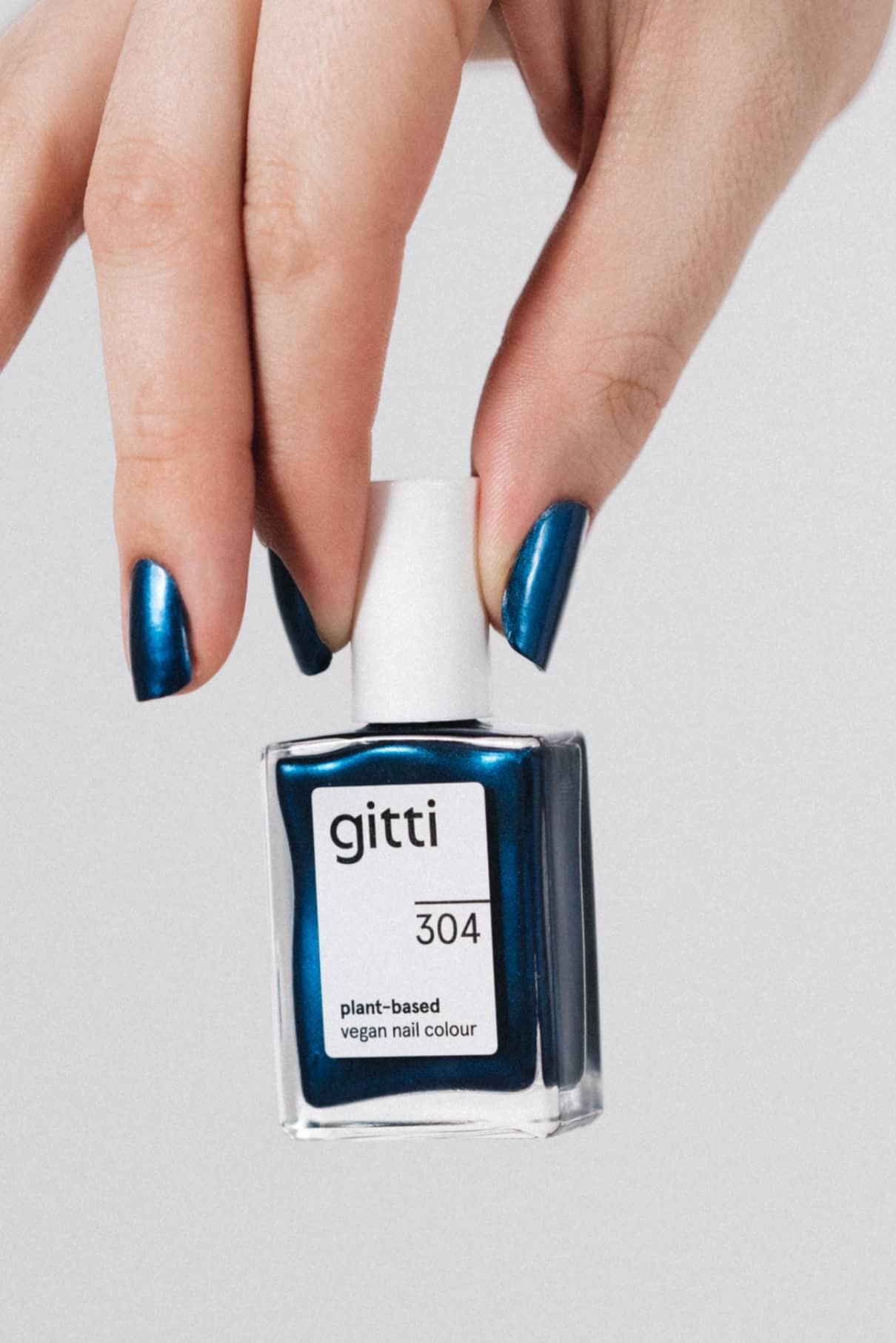 Eine Hand mit Metallic-Blauen Nägeln hält eine Flasche gitti no. 304 pflanzlicher veganer Nagelfarbe vor einem hellgrauen Hintergrund.