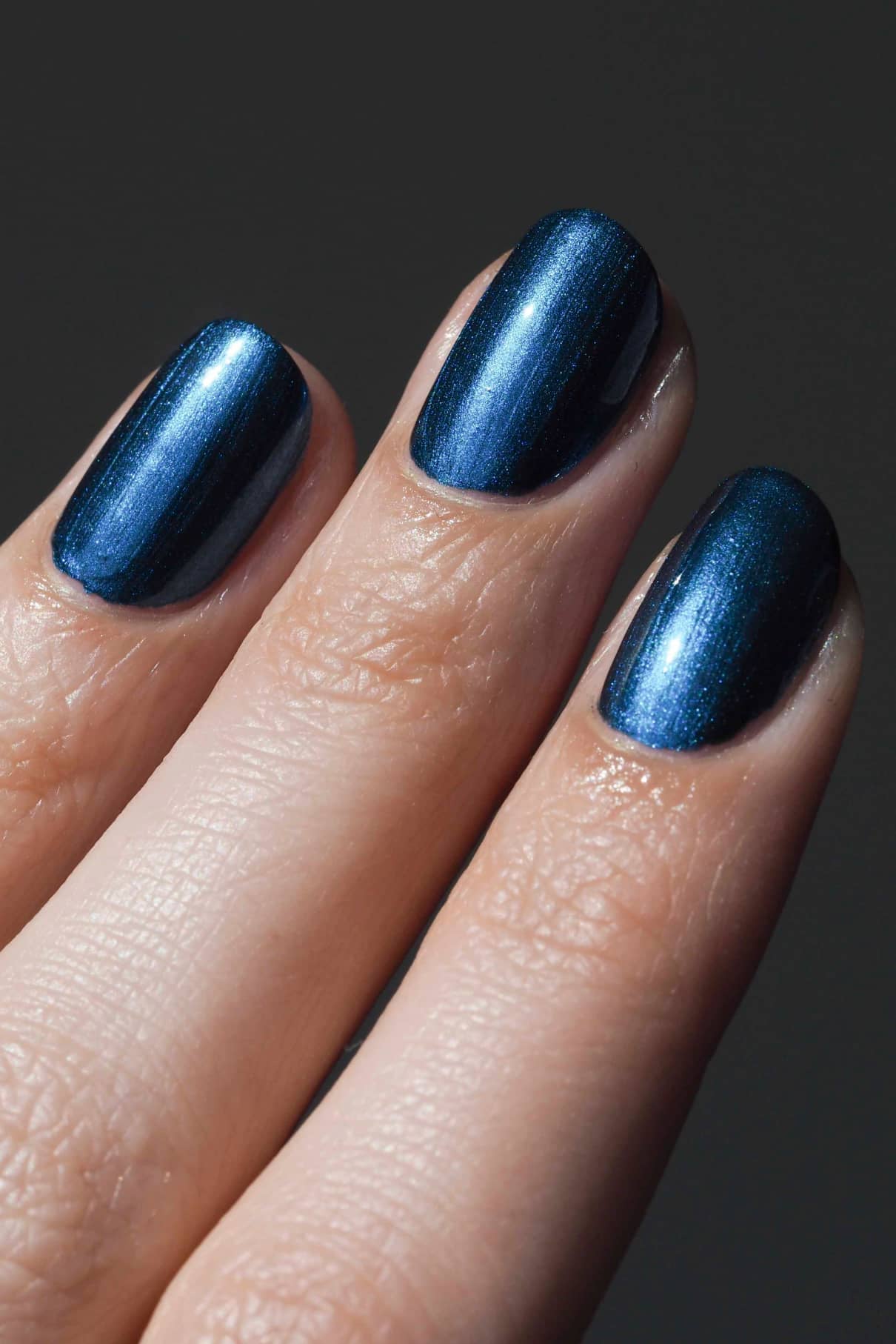 Nahaufnahme von drei Fingern mit kurzen, gut gepflegten Nägeln, lackiert mit gitti no. 304 Metallic-Blauem Nagellack vor dunklem Hintergrund.