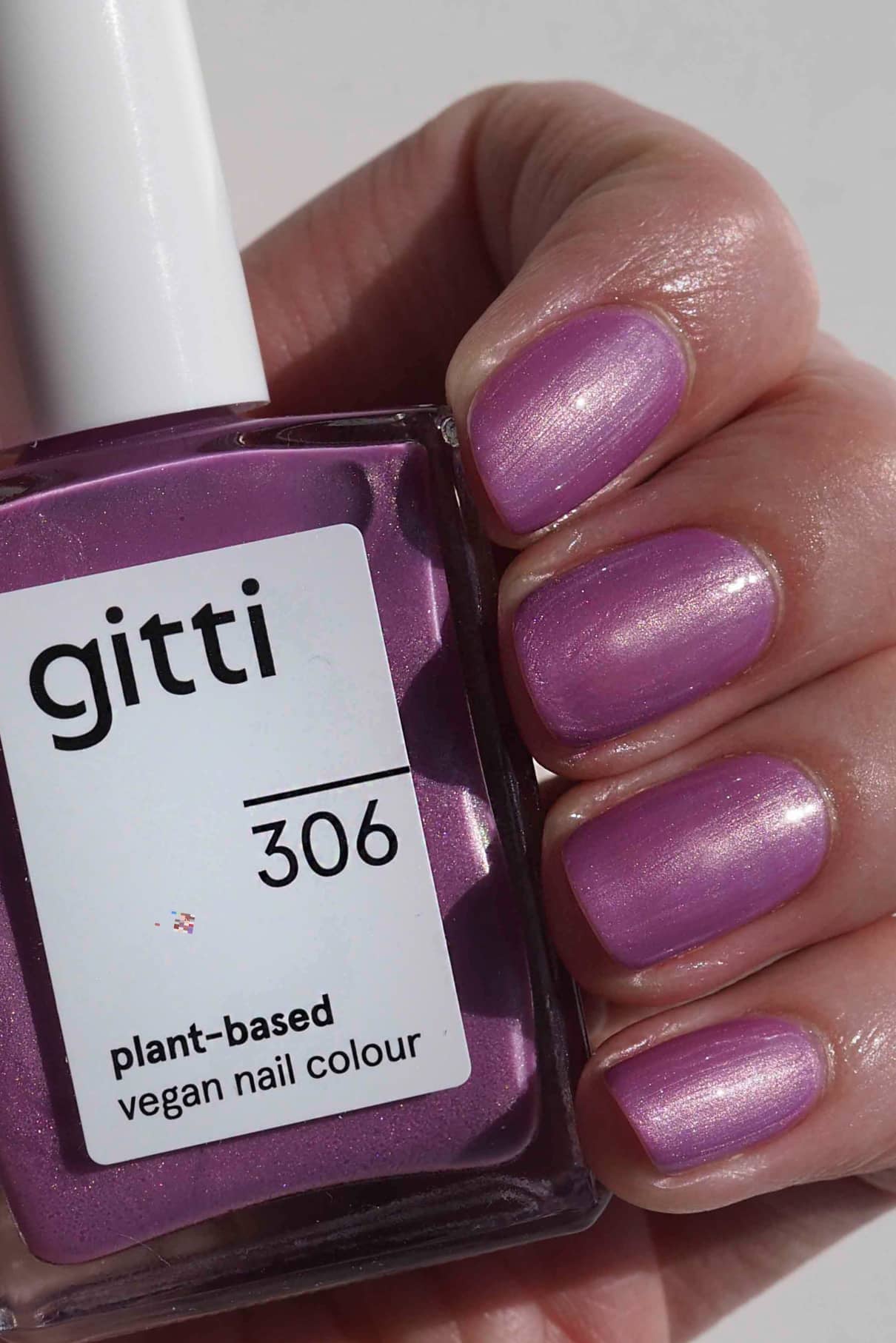 Eine Hand mit kurzen Nägeln, die mit schimmerndem Lavendellack mit violettem Unterton lackiert sind, hält eine Flasche mit gitti no. 306, einer pflanzlichen veganen Nagelfarbe in einem passenden Metallic-Mauve-Ton.