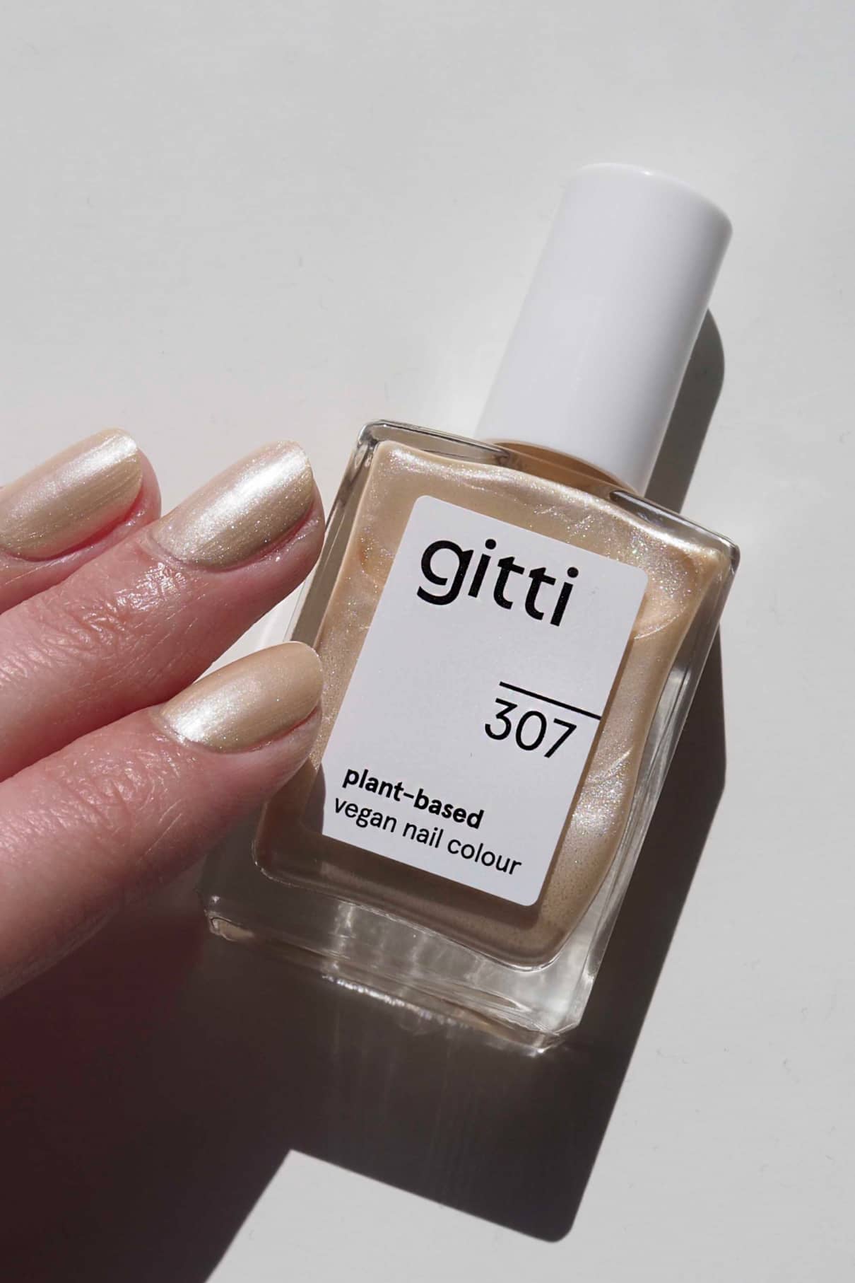 Eine Hand mit perlmuttbeigen Nägeln hält eine Flasche gitti no. 307, eine pflanzliche vegane Nagelfarbe in zartem Roségold-Creme-Metallic, vor einem weißen Hintergrund.