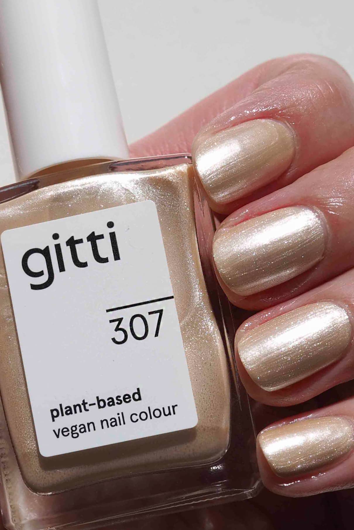 Eine Hand mit Nägeln, die mit gitti no. 307, einer sanft schimmernden, hellbeigen veganen Nagelfarbe auf Pflanzenbasis, hält das passende Fläschchen - ideal für Fans von zarten metallischen Cremetönen.