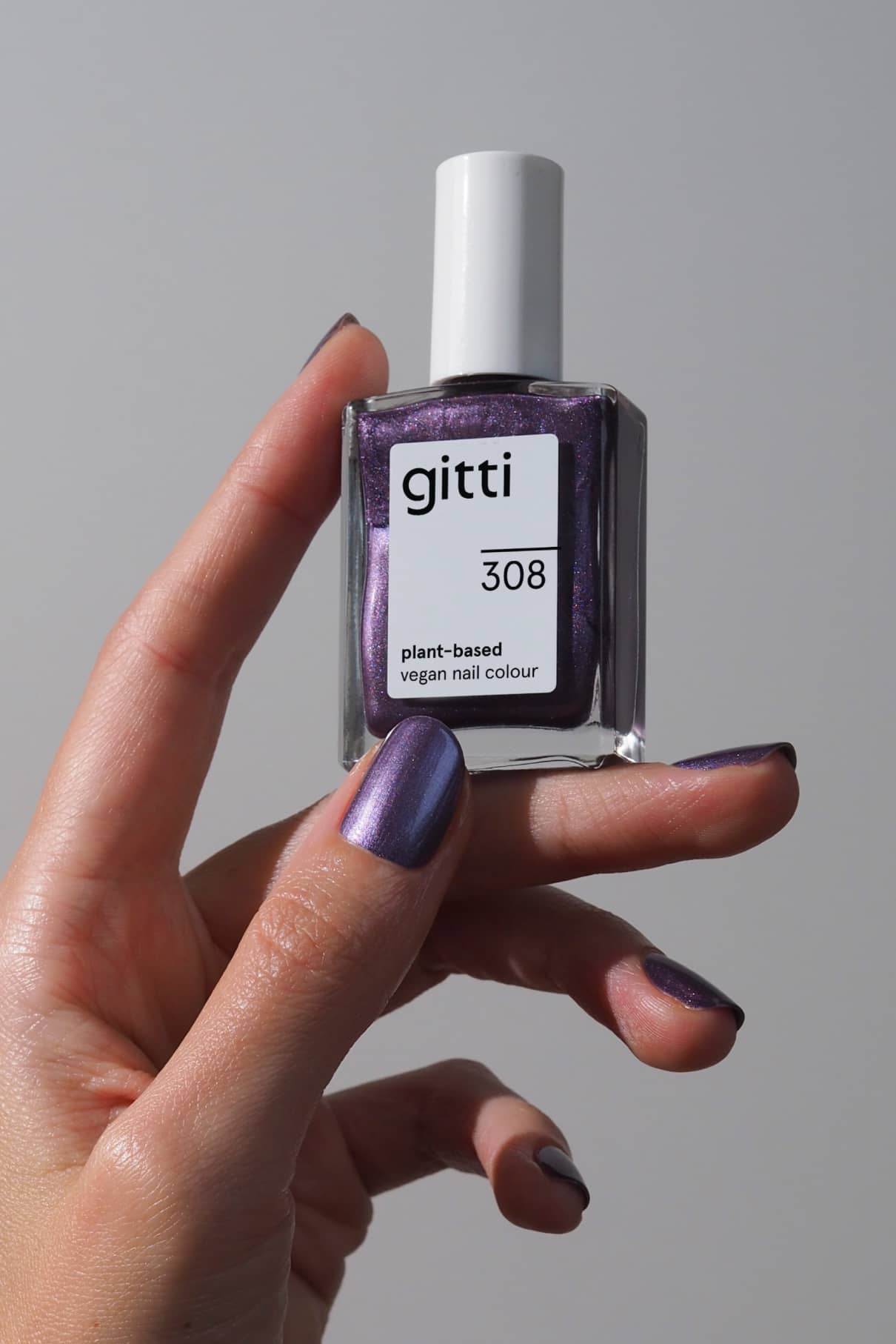 Eine Hand mit metallisch schimmernden lila Nägeln hält eine Flasche gitti no. 308 pflanzlichen, veganen Nagellack im Farbton Violettton vor einem schlichten, hellen Hintergrund.