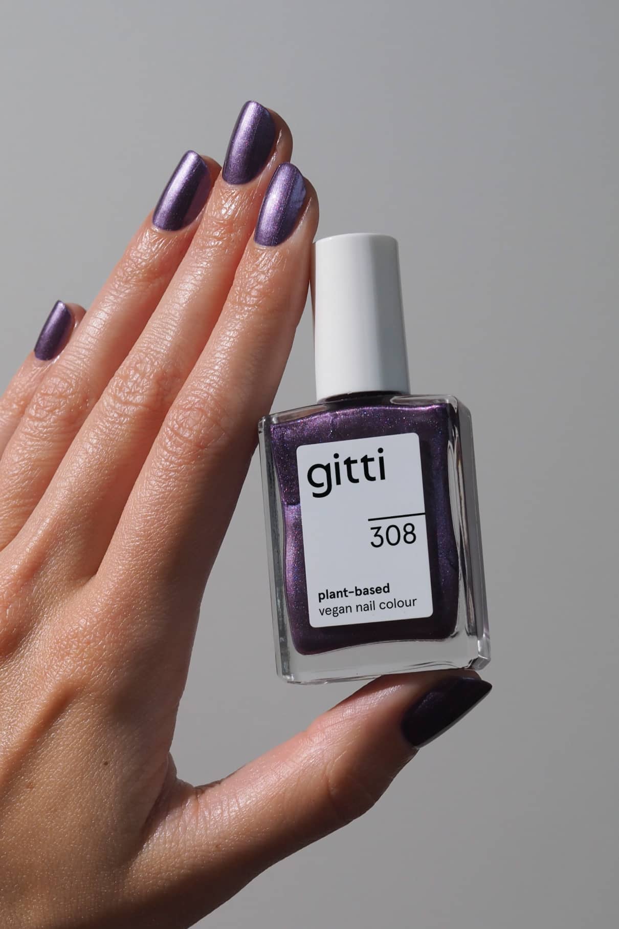 Eine Hand mit metallisch lila lackierten Nägeln in gitti no. 308 lackierten Nägeln hält das pflanzliche, vegane Nagelfarbfläschchen vor einem schlichten Hintergrund.