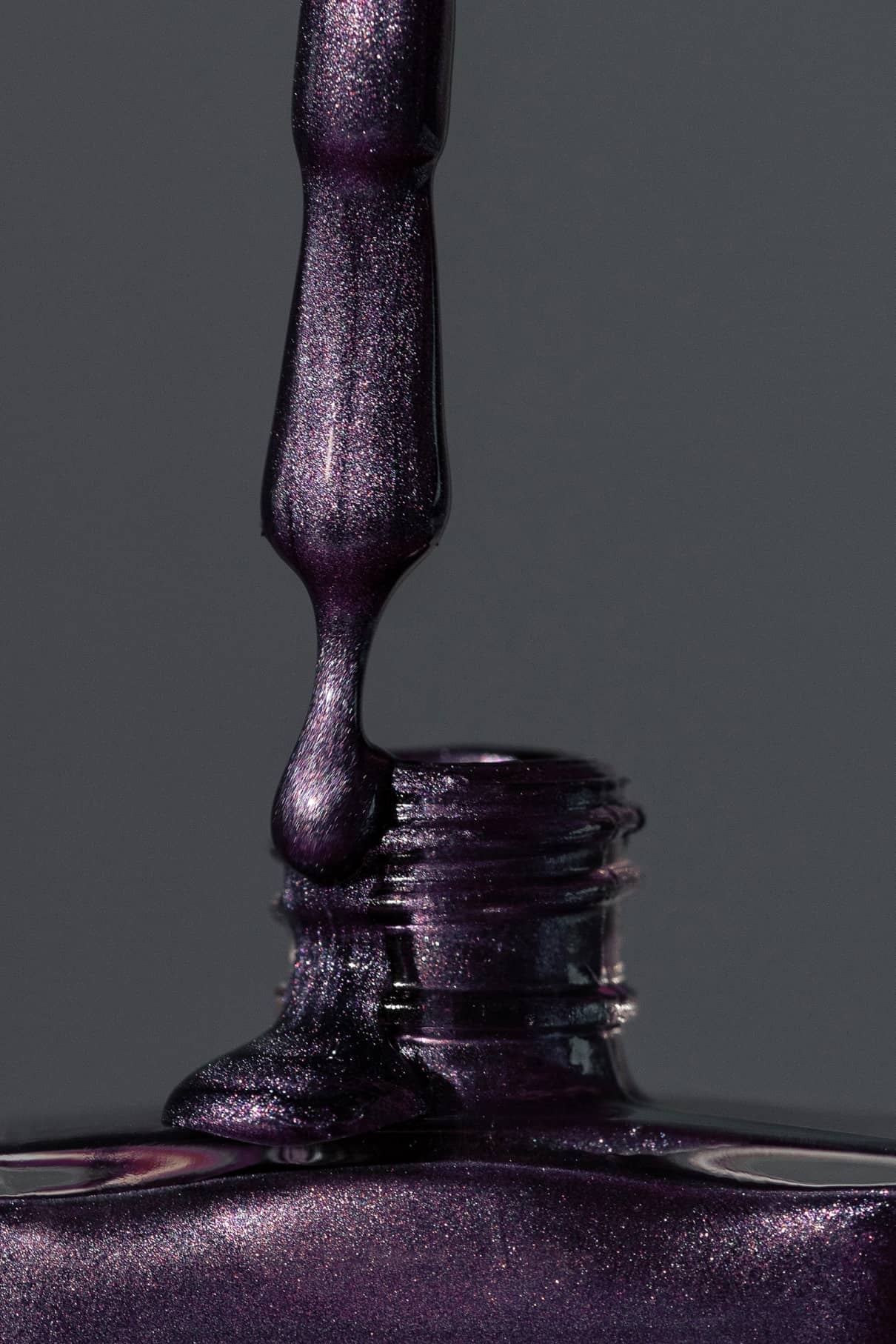 Eine Nahaufnahme eines Nagellackpinsels, der gitti no. 308 dunkelviolett schimmernder Metallic-Lack in eine offene Flasche vor einem grauen Hintergrund.