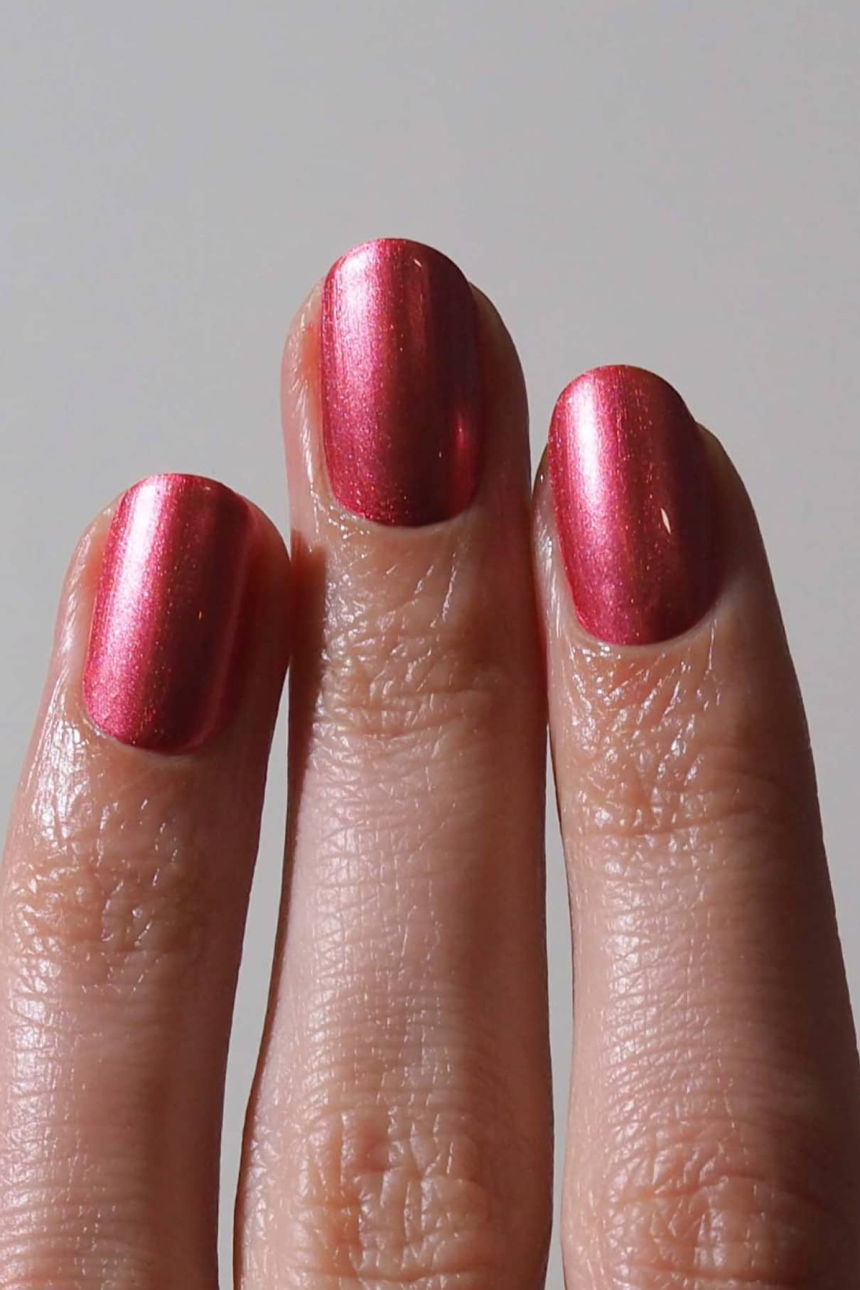 Drei Finger mit gitti no. 309, einem rötlich-pinkfarbenen Metallic-Nagellack, sind vor einem unifarbenen Hintergrund abgebildet.