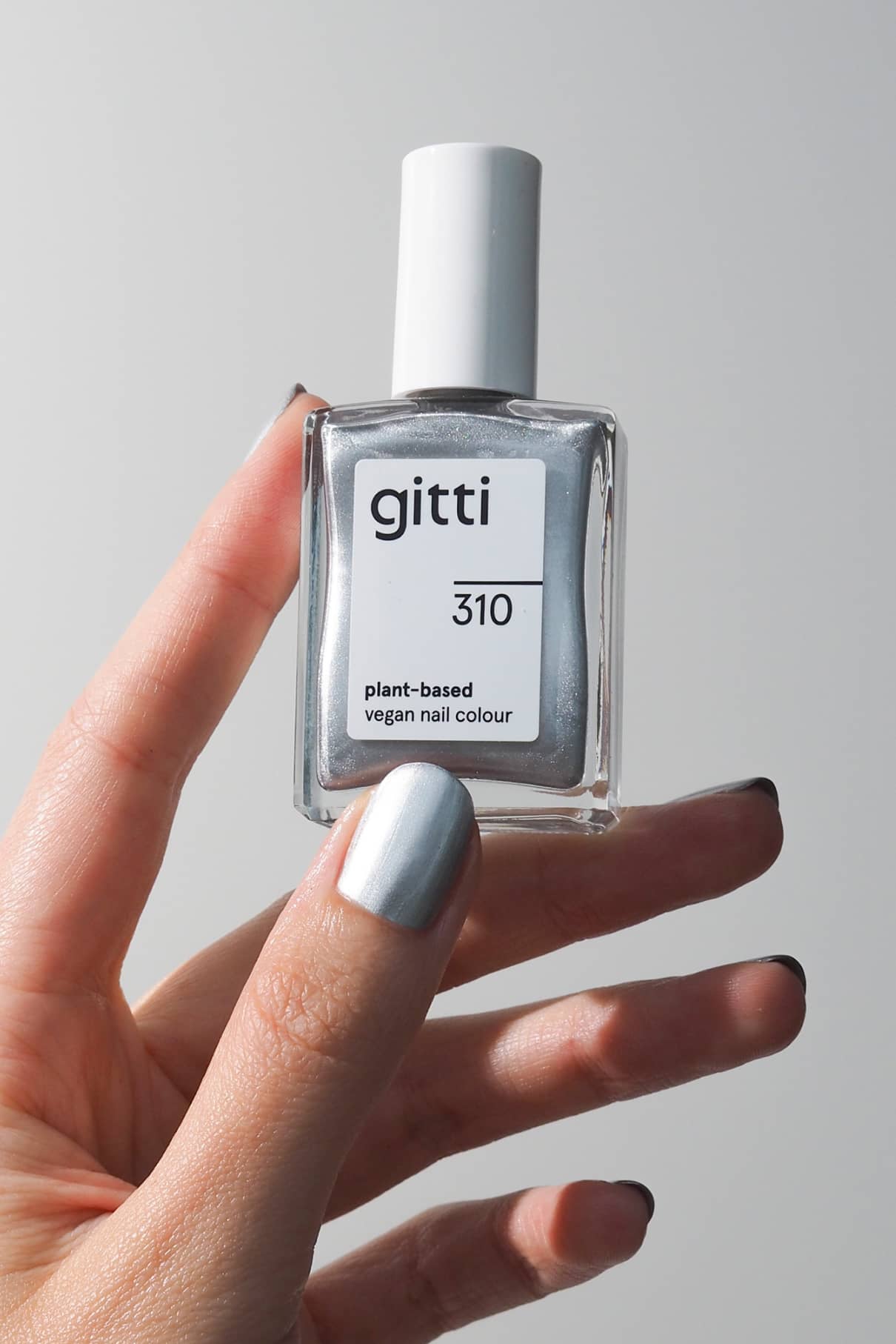 Eine Hand mit metallisch silber lackierten Nägeln, die einen auffälligen Chromeffekt aufweisen, hält eine Flasche gitti no. 310 pflanzlicher veganer Nagelfarbe vor einem schlichten Hintergrund.