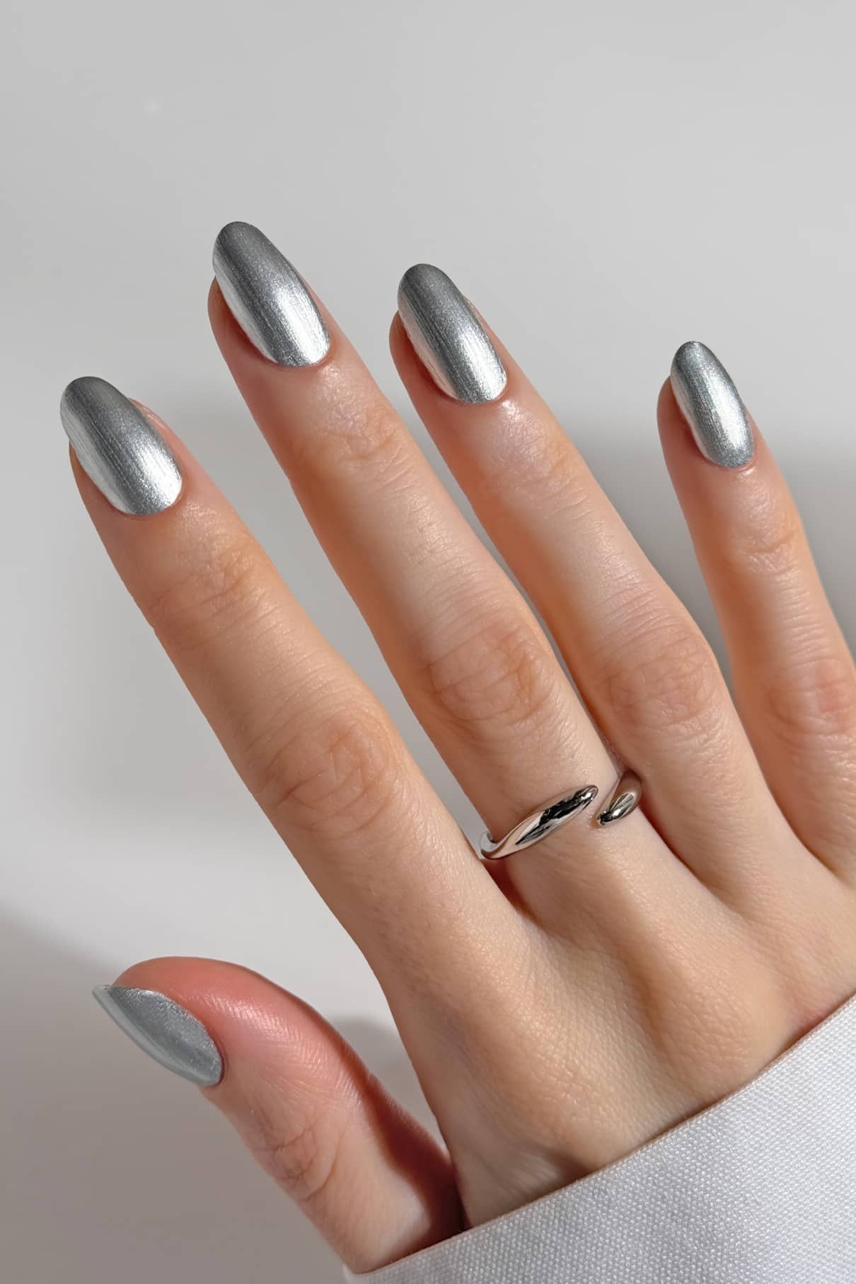 Eine Hand mit sauber manikürten gitti no. 310 Nägel in metallischem Silber mit dezentem Chrom-Effekt, mit einem schlichten silbernen Ring am Ringfinger, vor einem schlichten weißen Hintergrund.