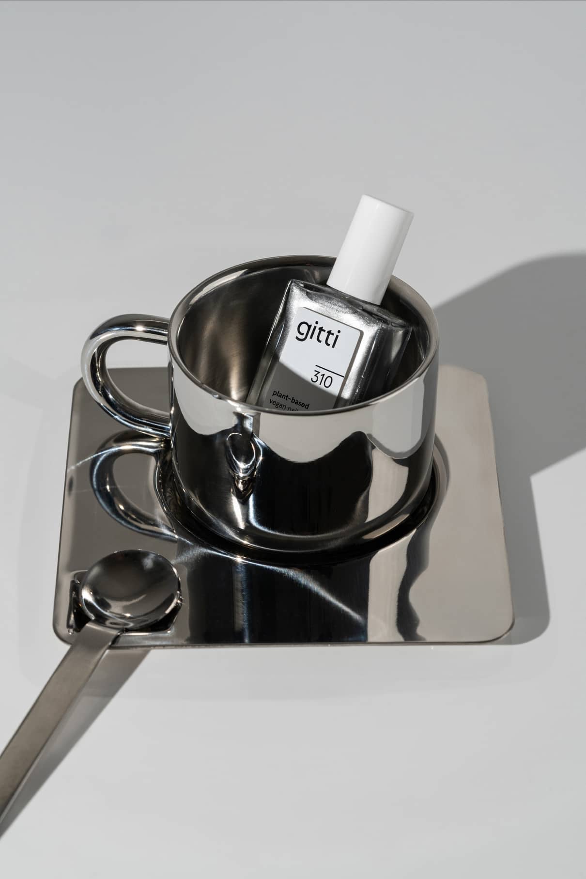 Eine Flasche gitti no. 310 Nagellack ist in einer reflektierenden Metalltasse auf einer passenden Untertasse untergebracht, die ihren Chrom-Effekt in metallischem Silber zeigt, mit einem Metalllöffel daneben auf einer weißen Oberfläche.