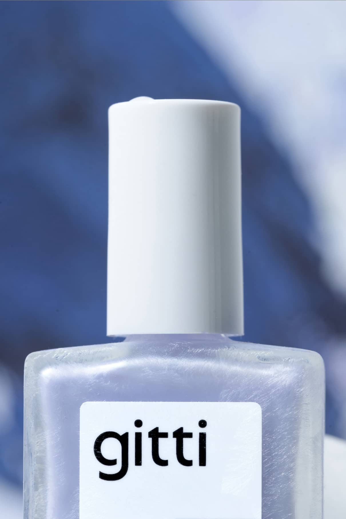 Eine Nahaufnahme einer Flasche gitti no. 311 Nagellack mit weißer Kappe und blassem Lila-Farbton mit dezentem Metallic-Finish vor einem unscharfen Hintergrund in kühlem Blau und Weiß.
