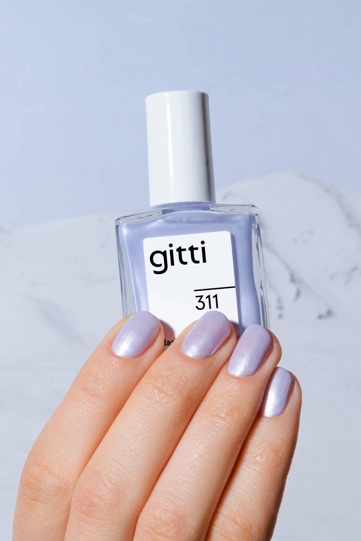Eine Hand mit kurzen, abgerundeten Nägeln, die mit schimmerndem, blasslila Lack mit Metallic-Finish lackiert sind, hält ein Fläschchen mit gitti no. 311 Nagelfarbe.