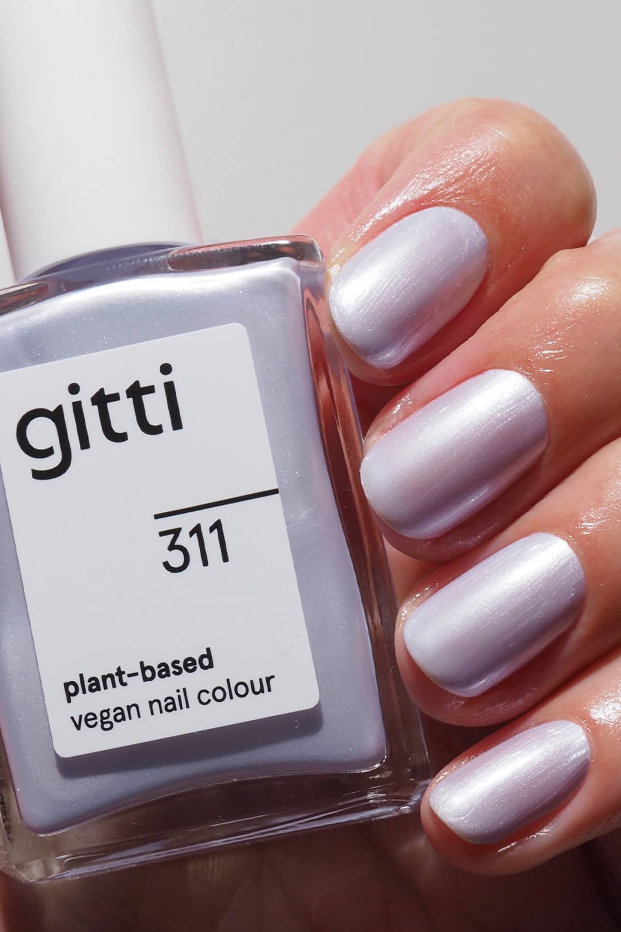 Eine Hand mit kurzen, in einem perlmuttfarbenen Lila-Ton lackierten Nägeln hält eine Flasche gitti no. 311, einer pflanzlichen veganen Nagelfarbe in einem kühlen blauen Metallic-Finish, das zum Nagelton passt.