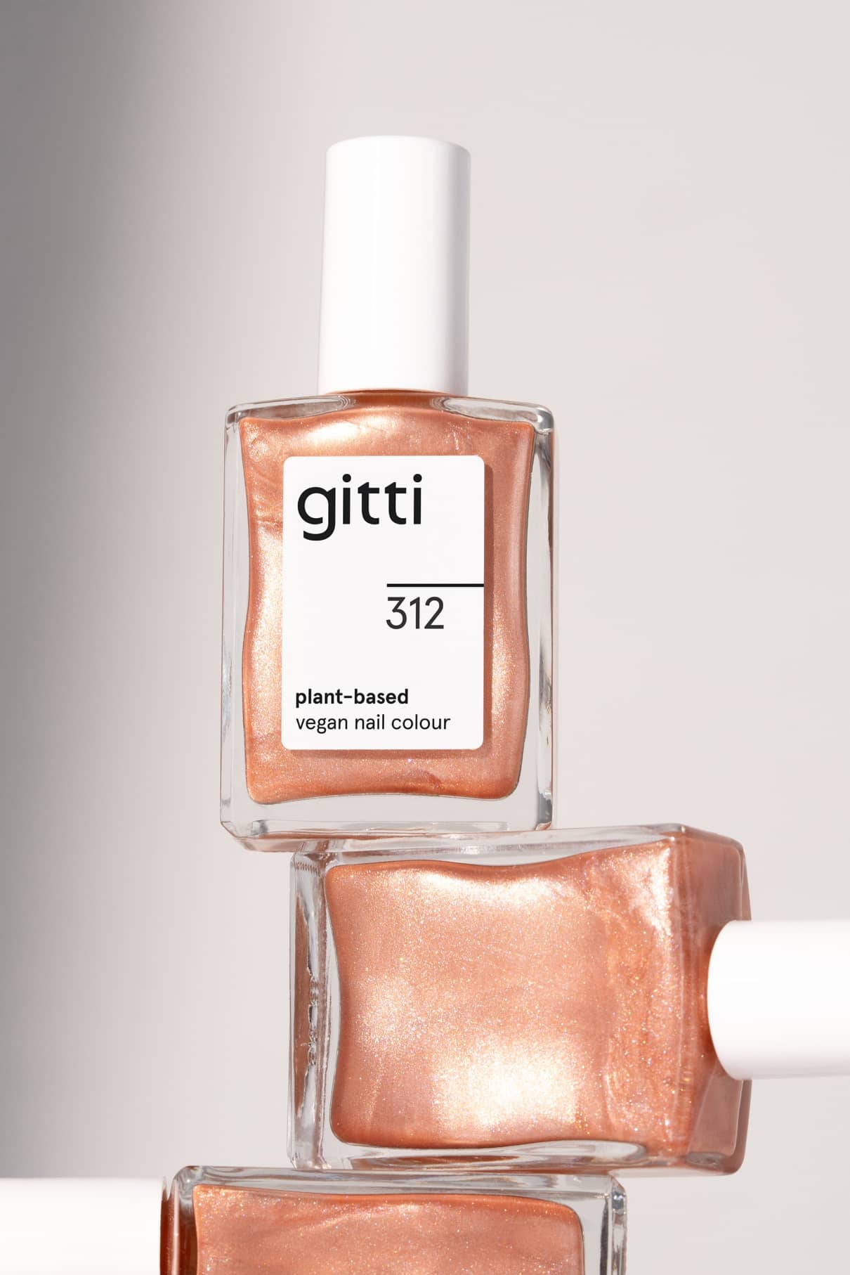 Drei Flaschen von gitti no. 312, ein veganer Nagellack auf pflanzlicher Basis in einem metallischen Apricot-Roségold-Ton, sind ordentlich vor einem neutralen Hintergrund gestapelt.