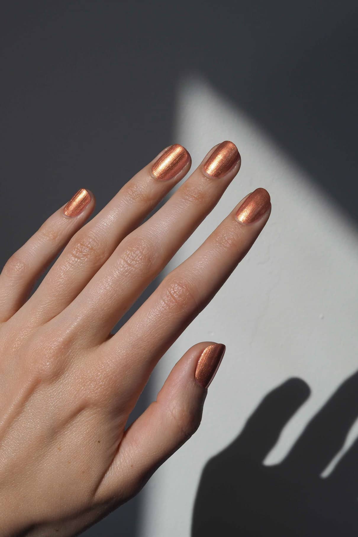 Eine Hand mit gitti no. 312 Metallic-Apricot-Nagellack ist vor einem weißen und dunkel schattierten Hintergrund abgebildet.