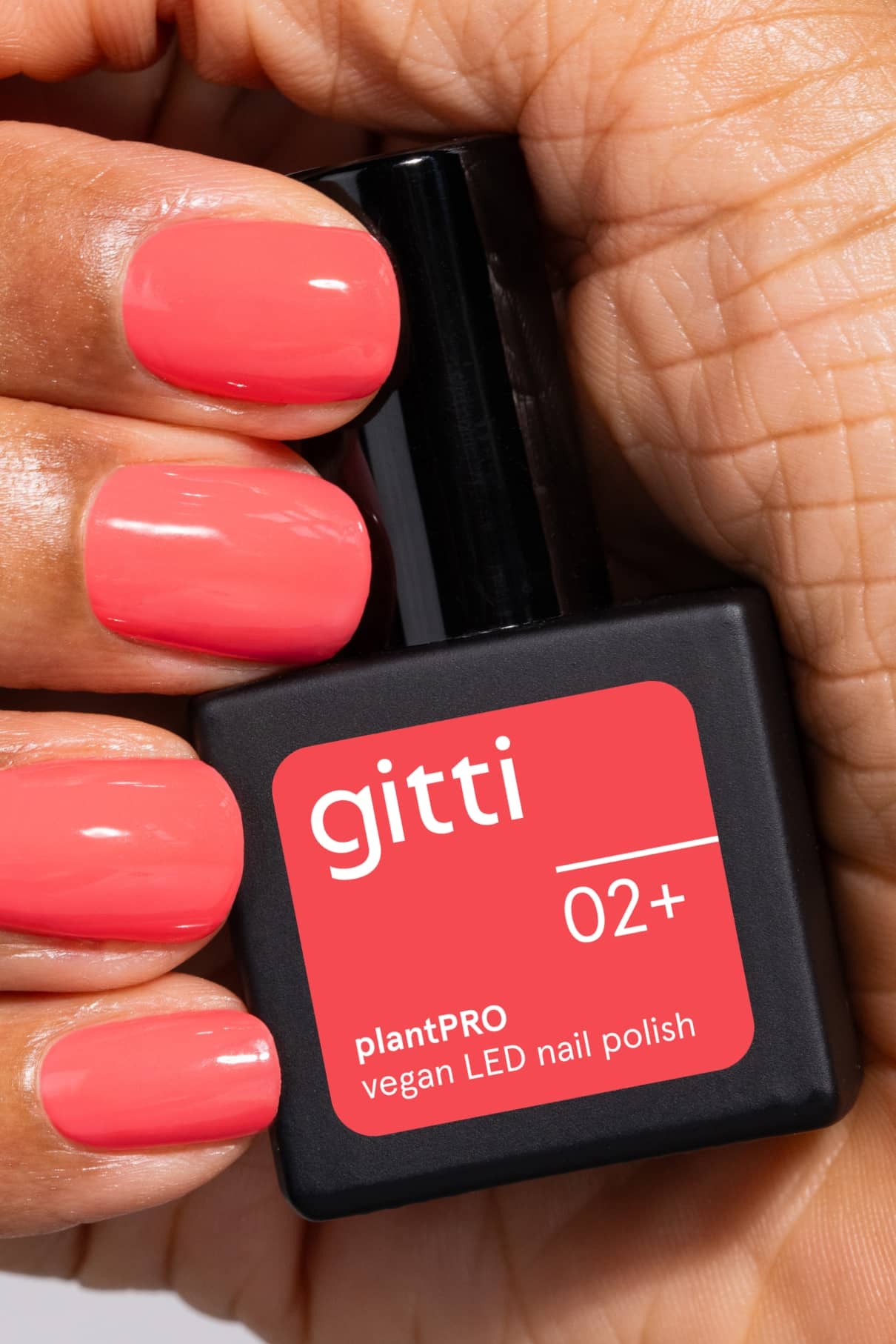 Eine Hand mit leuchtend korallfarbenen Nägeln und warmem Unterton hält eine schwarze Flasche mit der Aufschrift gitti no. 02+ plantPRO vegan LED Nagellack.