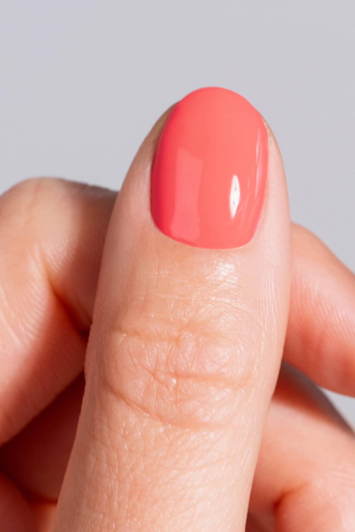 Eine Nahaufnahme eines Daumens mit dem gitti no. 02+ Nagellack - ein glatter, glänzender leuchtendes Korall-Ton mit einem wärmeren Unterton - vor einem neutralen Hintergrund.