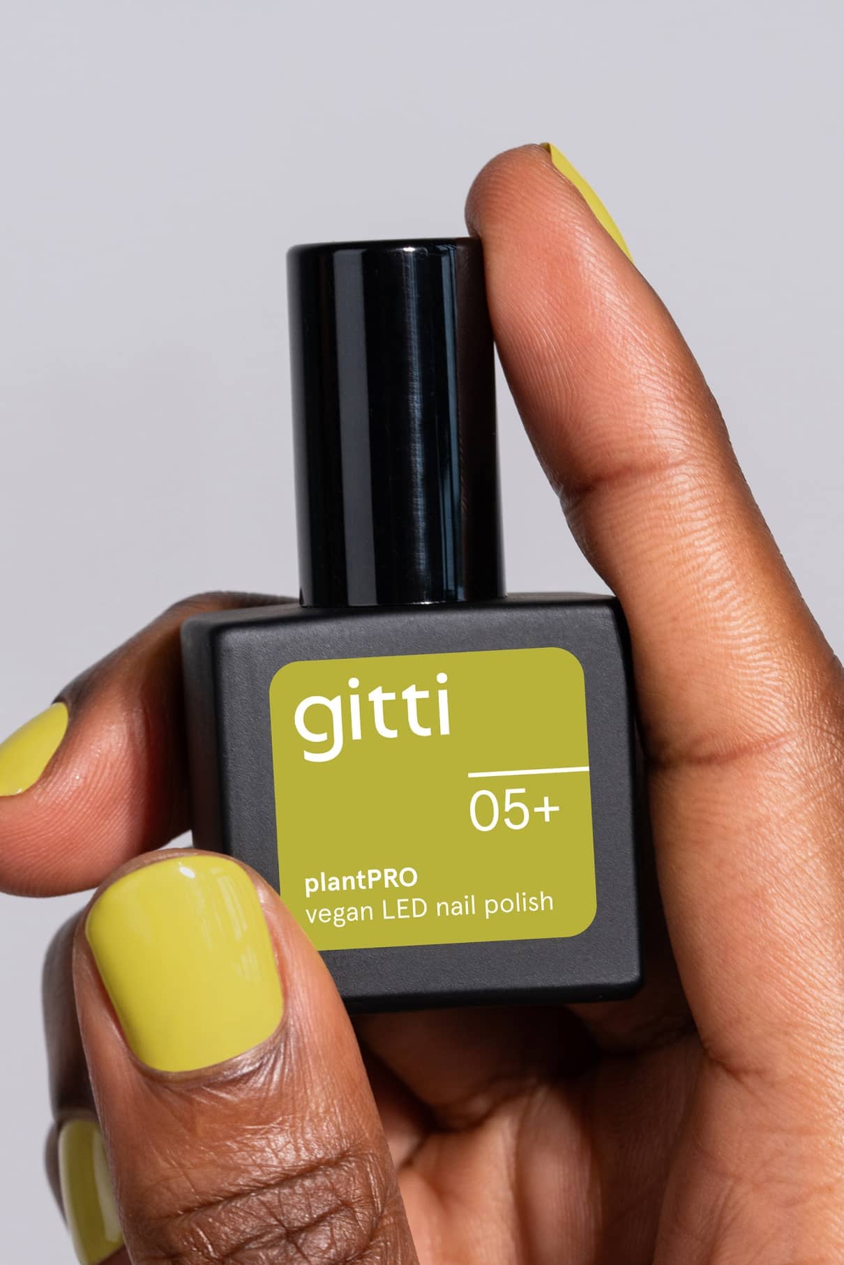 Eine Hand mit gelb-grün lackierten Nägeln hält eine viereckige Flasche mit gitti no. 05+ LED-Nagellack auf pflanzlicher Basis, einem modernen Grünton mit gelbem Unterton.