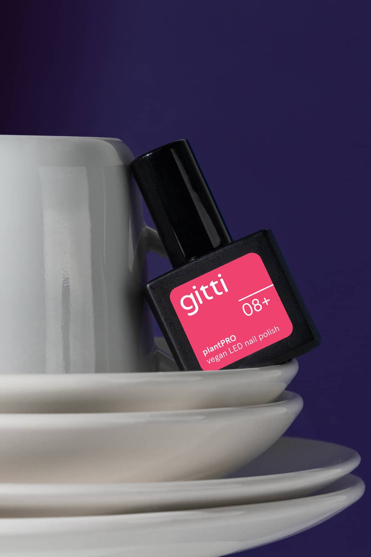 Ein weißes Tassen- und Untertassenset mit einer Flasche gitti no. 08+ veganer LED-Nagellack in leuchtendem Pink lehnt an der Tasse, alles vor einem lila Hintergrund.